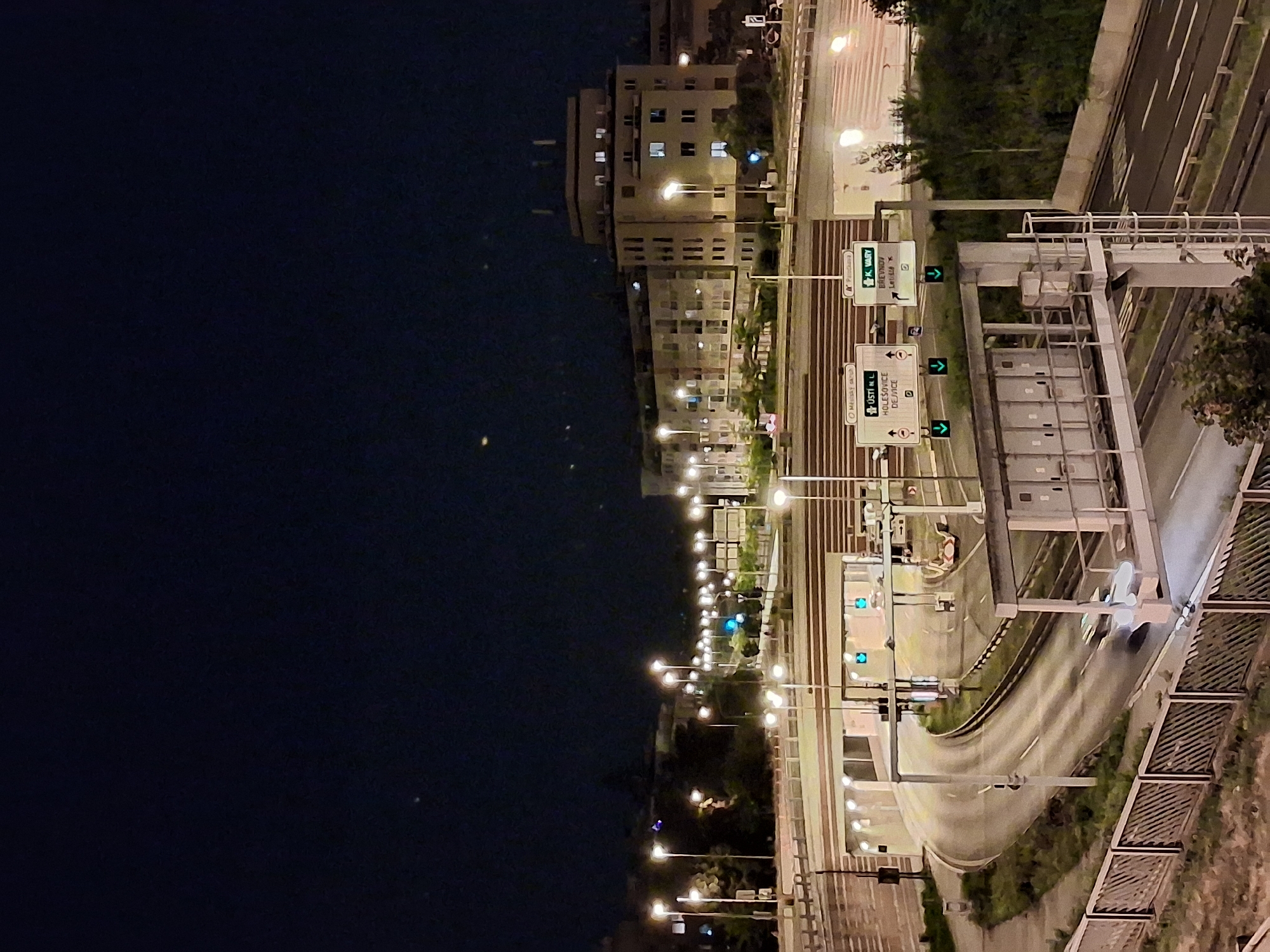 The image captures a nighttime view of a highway interchange in Prague, Czechia. Illuminated by streetlights and tunnel lights, the scene shows a multi-lane road curving around a tunnel entrance. Overhead signs indicate directions to "Ústí n. L." and "K. Vary," suggesting these are major routes. A few cars are visible on the road, one of which is a white vehicle moving through the interchange. Buildings line the background, with windows lit from within, indicating urban activity. The sky above is dark, suggesting it is late at night. The overall impression is of a busy, modern transportation hub operating under the cover of darkness.