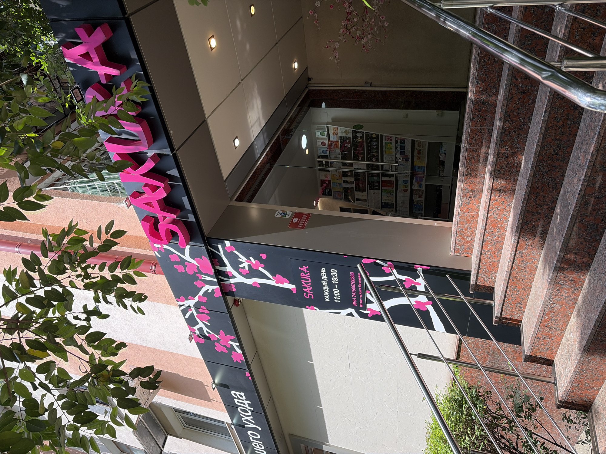 The image displays the sunlit exterior entrance of a retail establishment named "Sakura" in Sochi, Russia. A set of five reddish-brown speckled granite steps, equipped with stainless steel handrails on both sides, ascends to a glass-paneled double door entrance. The building's facade features a prominent dark grey sign adorned with a white cherry blossom (sakura) tree motif and pink flowers. Above the entrance, the word "SAKURA" is rendered in large, three-dimensional pink letters. A vertical dark sign to the immediate left of the entrance reiterates "SAKURA" in English, followed by Russian text indicating operating hours ("КАЖДЫЙ ДЕНЬ 11:00 – 19:30" - Every day 11:00 – 19:30) and legal entity information ("ИНН 741506700388 ИП Вагина Юлия Евгеньевна"). The upper section of the building exhibits alternating light peach and white horizontal stripes. Green leafy branches of an overhead tree partially obscure the top of the facade, casting shadows. A dense green bush is visible to the left of the stairs, behind the railing. Through the glass doors, the interior of the store reveals shelves stocked with various products. The scene is devoid of people.