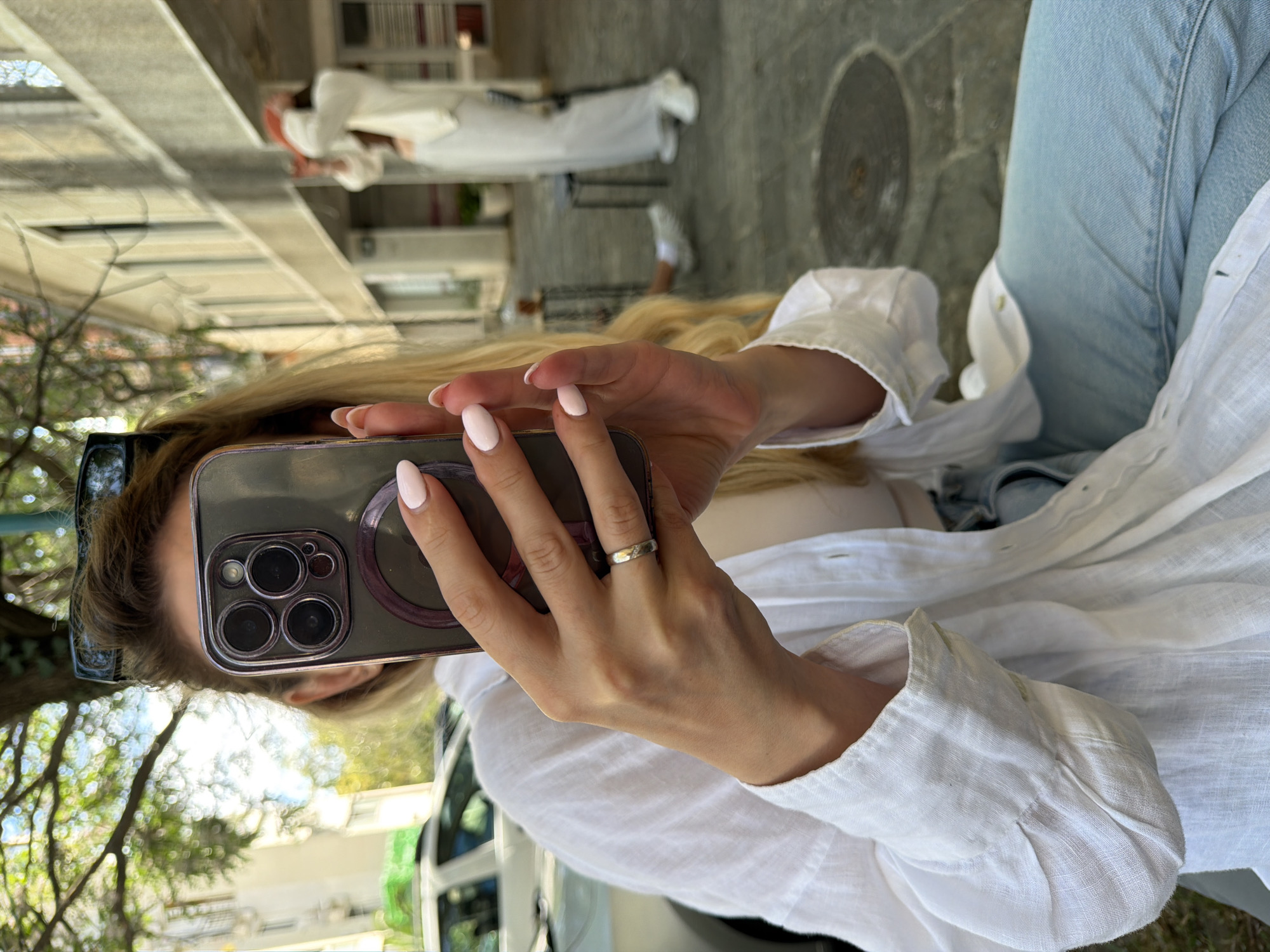A woman is holding a phone, a gray model with a ring on the back, to take a picture. She wears a white button-down linen shirt, jeans, and a silver ring on her right ring finger. She has long blonde hair, partially obscuring a pair of sunglasses.
In the background, slightly blurred, a person in a long white robe and headdress walks across a cobblestone street. A building with a beige facade and white-framed windows is also visible.
The location is likely in Budva, Montenegro, evidenced by the architecture, the cobblestone streets, and the style of dress of the individuals.