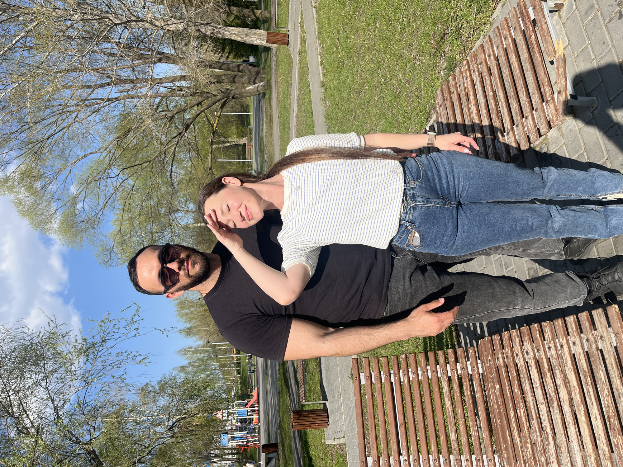 A couple, a man and a woman, are posing for a photo while sitting on a wooden bench in a park-like setting in Ulyanovsk, Russia. The woman, wearing a horizontally striped shirt and jeans, is leaning back on the man who is wearing a black t-shirt and sunglasses. Her hand is raised to her forehead. The man is looking toward the camera with his arm around the woman. They are positioned on a worn, brown wooden bench on top of a paved area. In the background there is green grass and trees. A playground is visible in the lower left corner of the image. The sky is blue with scattered clouds.