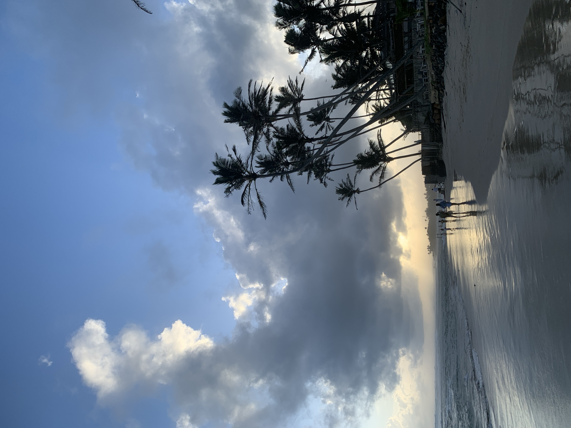 This image depicts a beach scene in Kathaluwa, Sri Lanka, with a dramatic sky featuring a mix of dark, heavy clouds and patches of bright sunlight. The ocean on the left shows gentle waves crashing onto a wet, sandy shore that reflects the sky and palm trees. A line of silhouetted palm trees leans towards the sea on the right side of the image, with a building structure partially visible behind them. Several people are walking along the waterline, their figures silhouetted against the bright reflections on the sand. The overall atmosphere is that of a tropical beach at either sunrise or sunset, with a sense of impending or passing weather.