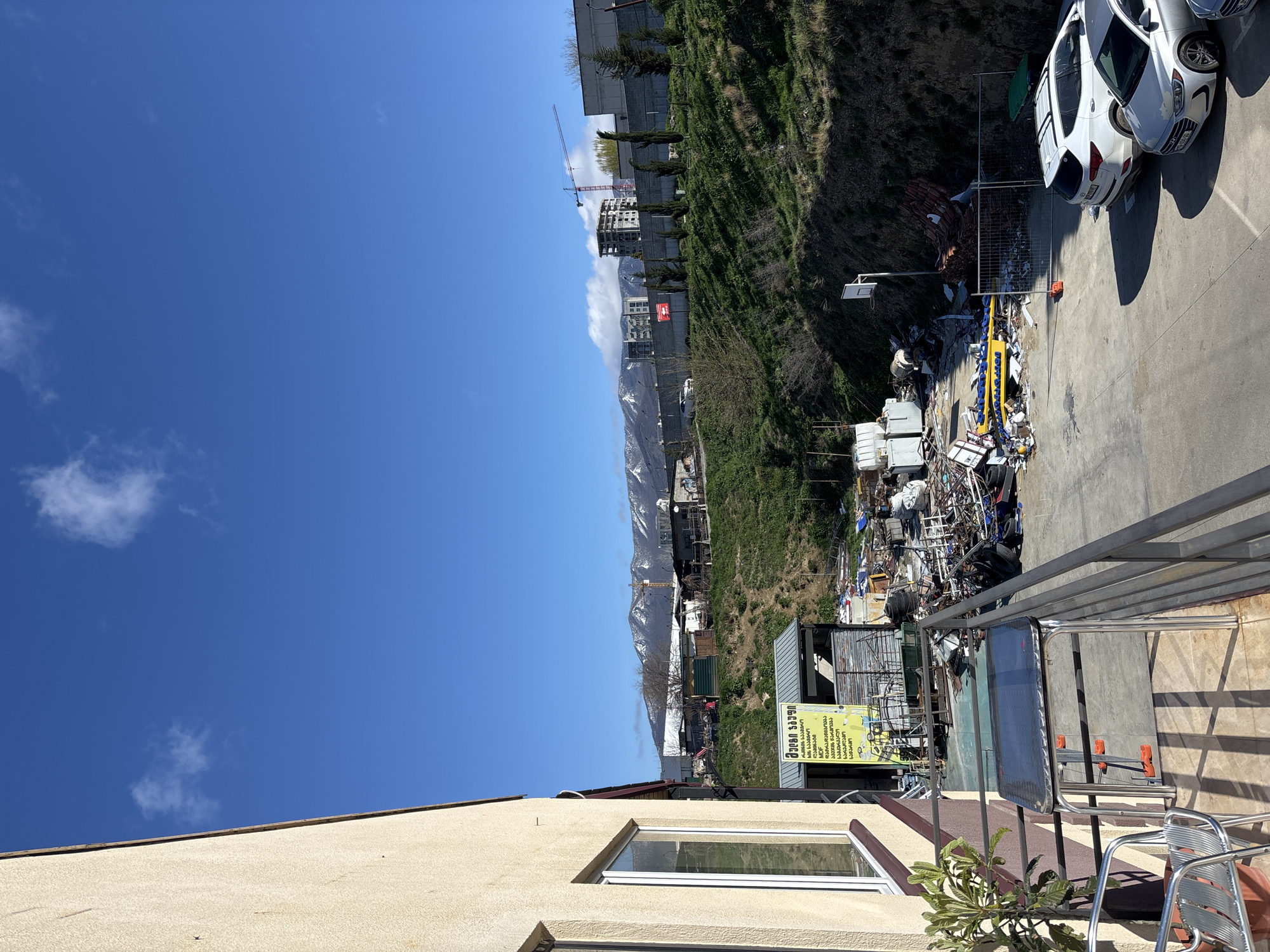 The image depicts a scene from what appears to be a balcony or elevated platform overlooking a cluttered area possibly located on the outskirts of Tbilisi, Georgia. The weather is clear and sunny.
In the foreground, there is a partial view of a building with a window, and a balcony with metal railings, a table, and chairs.
In the midground, a large pile of debris and scrap materials is visible, possibly indicating a construction or demolition site. There are also vehicles parked nearby, including white cars. Behind this area, a grassy hillside rises up, with sparse vegetation and some structures built into the hillside.
In the background, a mountain range is visible, with some peaks covered in snow. A construction crane suggests ongoing development in the area. The sky is clear and blue, with a few scattered clouds.
The image does not contain any people. The overall impression is one of a transitional urban environment, with construction and development occurring near a more natural landscape.