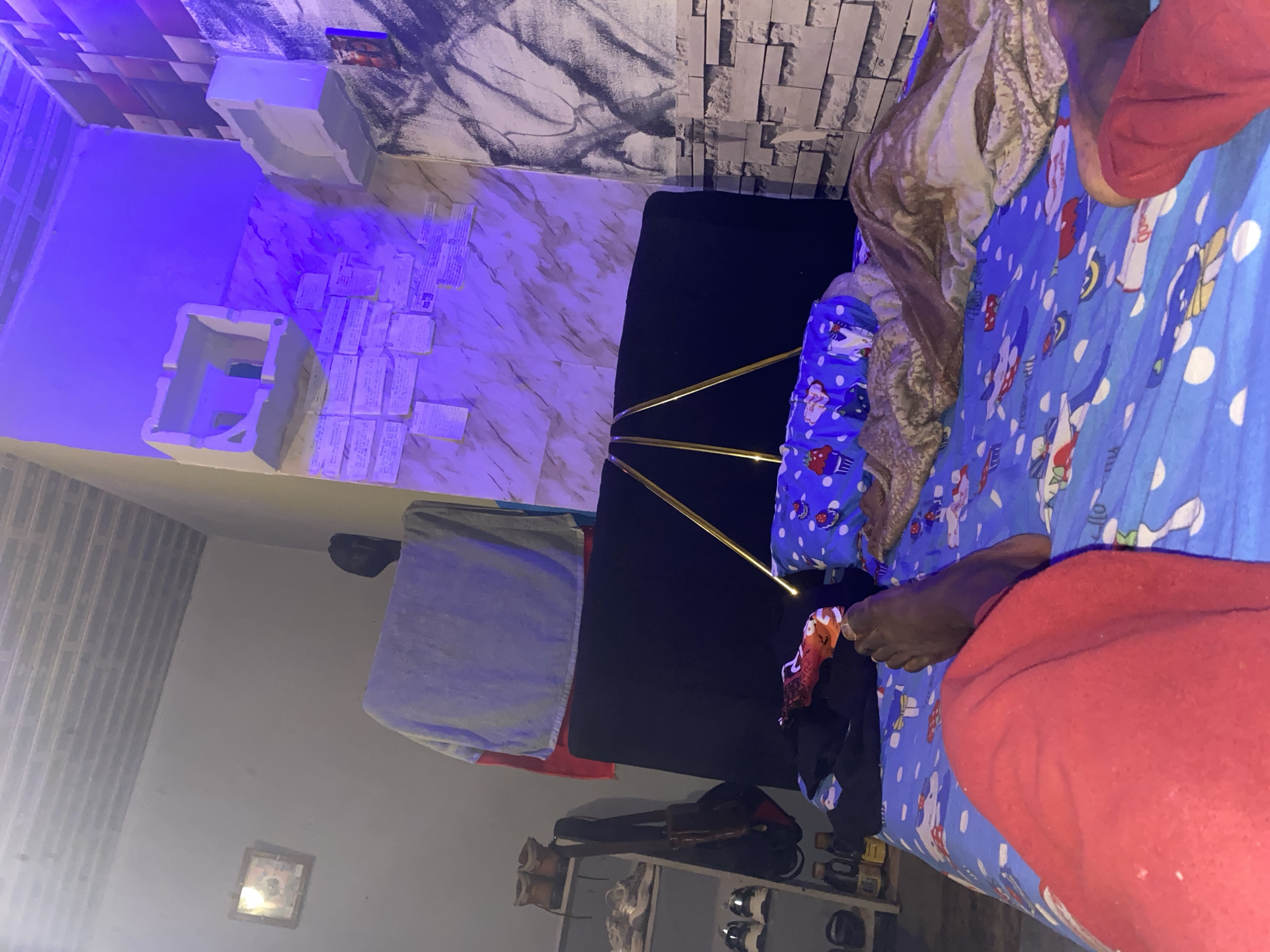 A low-angle, indoor shot captures a person lying on a bed in Kabong, Nigeria. The room is dimly lit with blue and purple ambient lighting. The bed is covered with blue sheets featuring cartoon characters, and a portion of the person's red clothing is visible. A pillow with a cupcake design rests on the bed, partially covered by a brown blanket. The background shows a headboard, shelves with shoes, a bag hanging on a rack, and a wall with a textured stone pattern and what appears to be a mural. The overall impression is of a cluttered but lived-in bedroom.