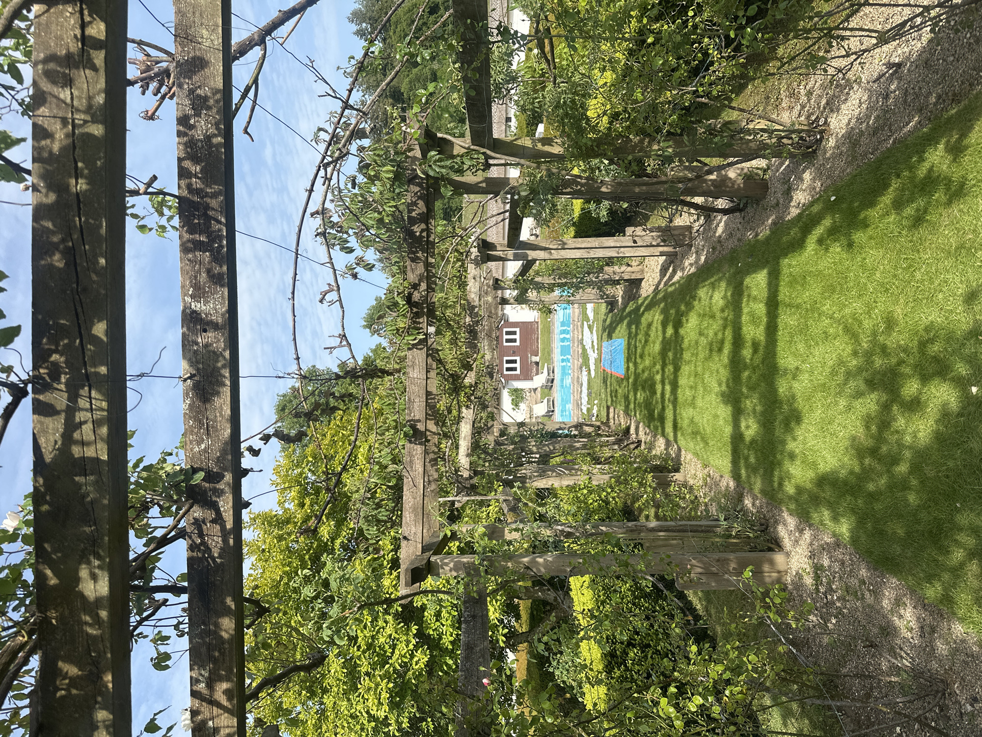 The image shows a garden path in Unknown, Unknown. The path is lined with a wooden pergola covered in foliage, creating a shaded tunnel. The path is made of gravel, and lush green grass borders it on the right. In the distance, a building with a swimming pool can be seen. A blue tarp or cover is visible near the pool. The sky is blue with some wispy clouds visible through the pergola.