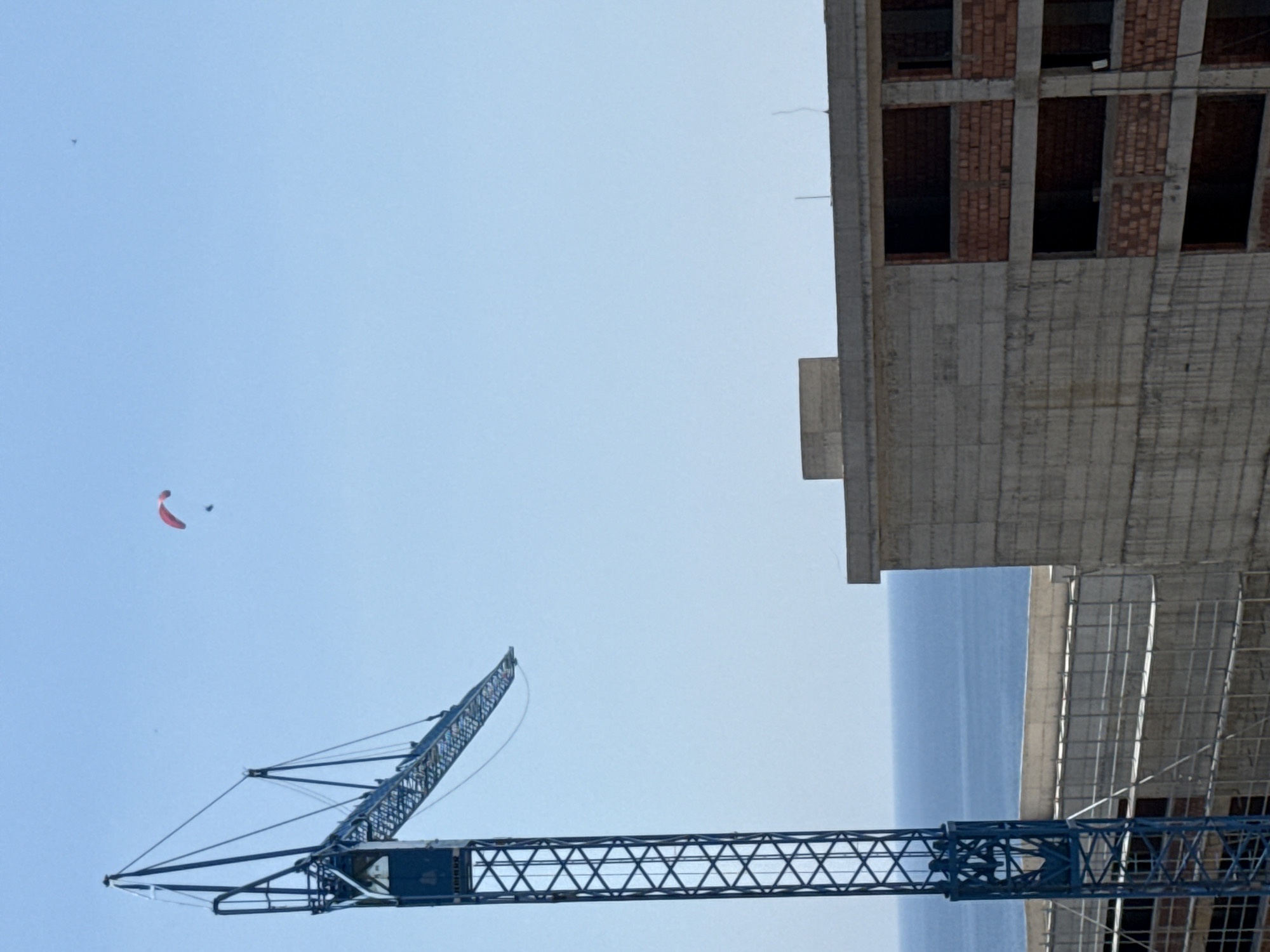 This media file depicts an outdoor scene dominated by a clear blue sky and a large construction site. A prominent feature is a dark blue construction crane, its latticework structure extending across the lower portion of the frame. In the upper right, the partially constructed facade of a building is visible. This building shows concrete elements and rows of red bricks, with dark openings suggesting windows or doorways.
In the sky, a small, red object, likely a paraglider or a kite, is suspended. A smaller, dark speck is also visible near it, which could be another object or a bird. The overall impression is one of ongoing construction in what appears to be a coastal area, as a body of water is visible in the background between the construction elements. The lighting suggests it is daytime, with the bright sky indicating fair weather. The scene evokes a sense of industrial activity and potential leisure or recreation with the presence of the paraglider.