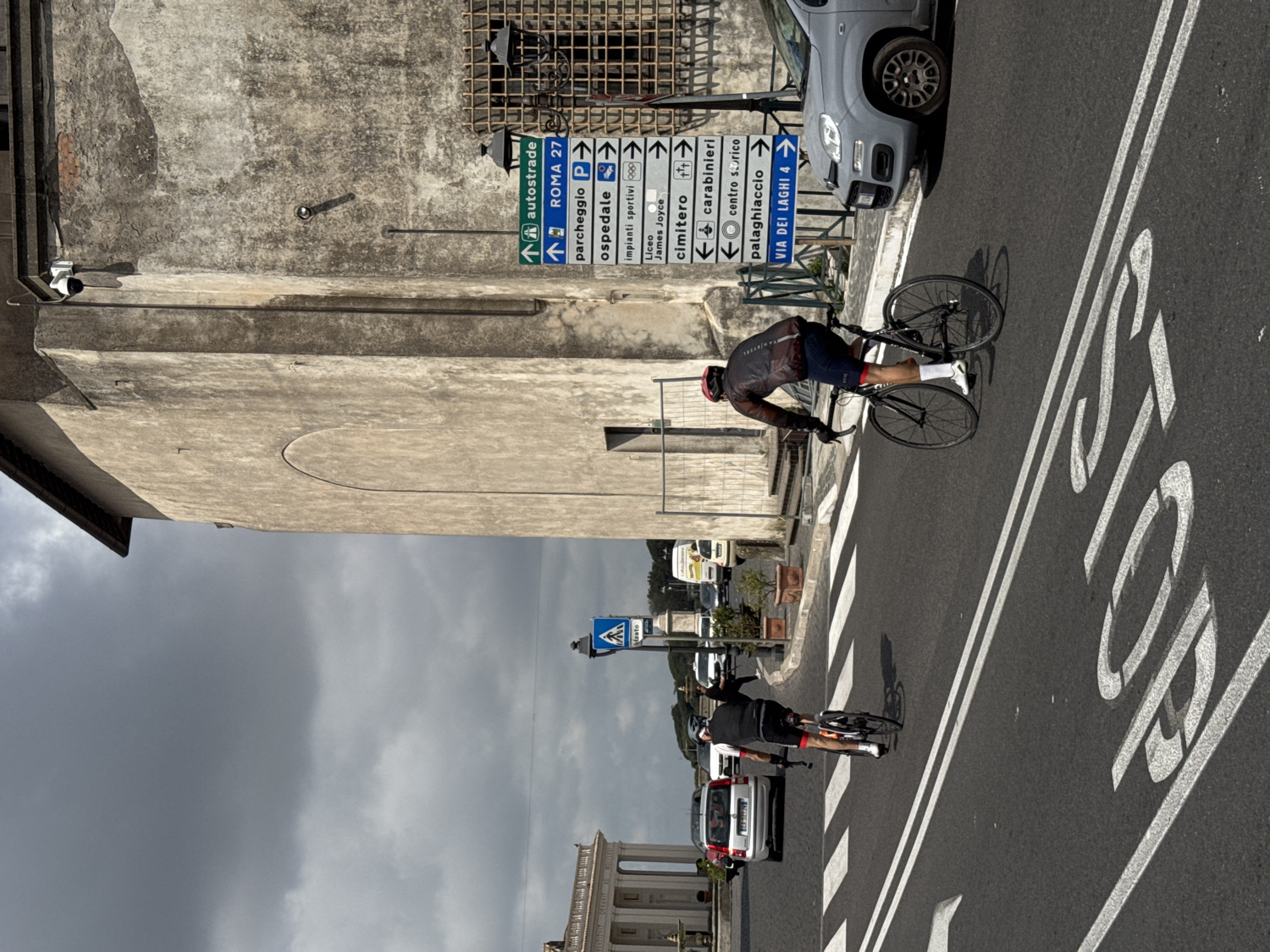 Two cyclists are riding on a paved road under an overcast sky in Ariccia, Italy. The scene captures a moment during daytime, possibly late morning or early afternoon, given the ambient light.
In the foreground, a cyclist wearing a dark and red cycling jersey, dark shorts, a red helmet, and white socks is positioned on a road bike, appearing to lean forward over the handlebars. Behind them, another cyclist in dark attire is also on a road bike. Both are on the right side of the road, which features prominent white road markings including a crosswalk and the word "STOP" painted on the asphalt.
To the right, a grey Fiat Panda car is parked alongside the curb. On the left side of the road, a large, weathered, light-colored building with a distinctive arched indention on its facade dominates the midground. Adjacent to this building, a multi-directional road sign displays various destinations in Italian, such as "↑ autostrade," "↑ ROMA 27," "parcheggio P," "ospedale," "impianti sportivi," "Liceo James Joyce," "cimitero +++," "← carabinieri," "← centro storico," "palaghiaccio →," and "VIA DEI LAGHI 4." Other buildings and street furniture are visible further down the road and in the background under the gray sky.