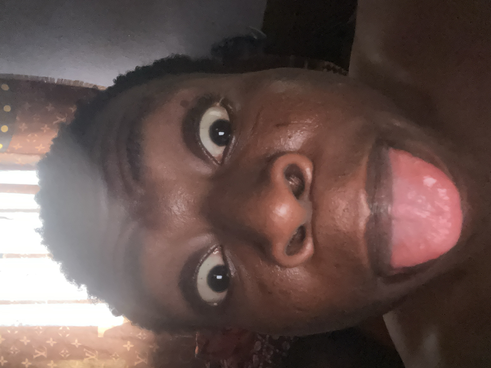 A close-up, rotated view of a dark-skinned person with their tongue extended. The person has wide eyes and short, curly hair. The background appears to include a patterned surface. The city and specific location are unknown.