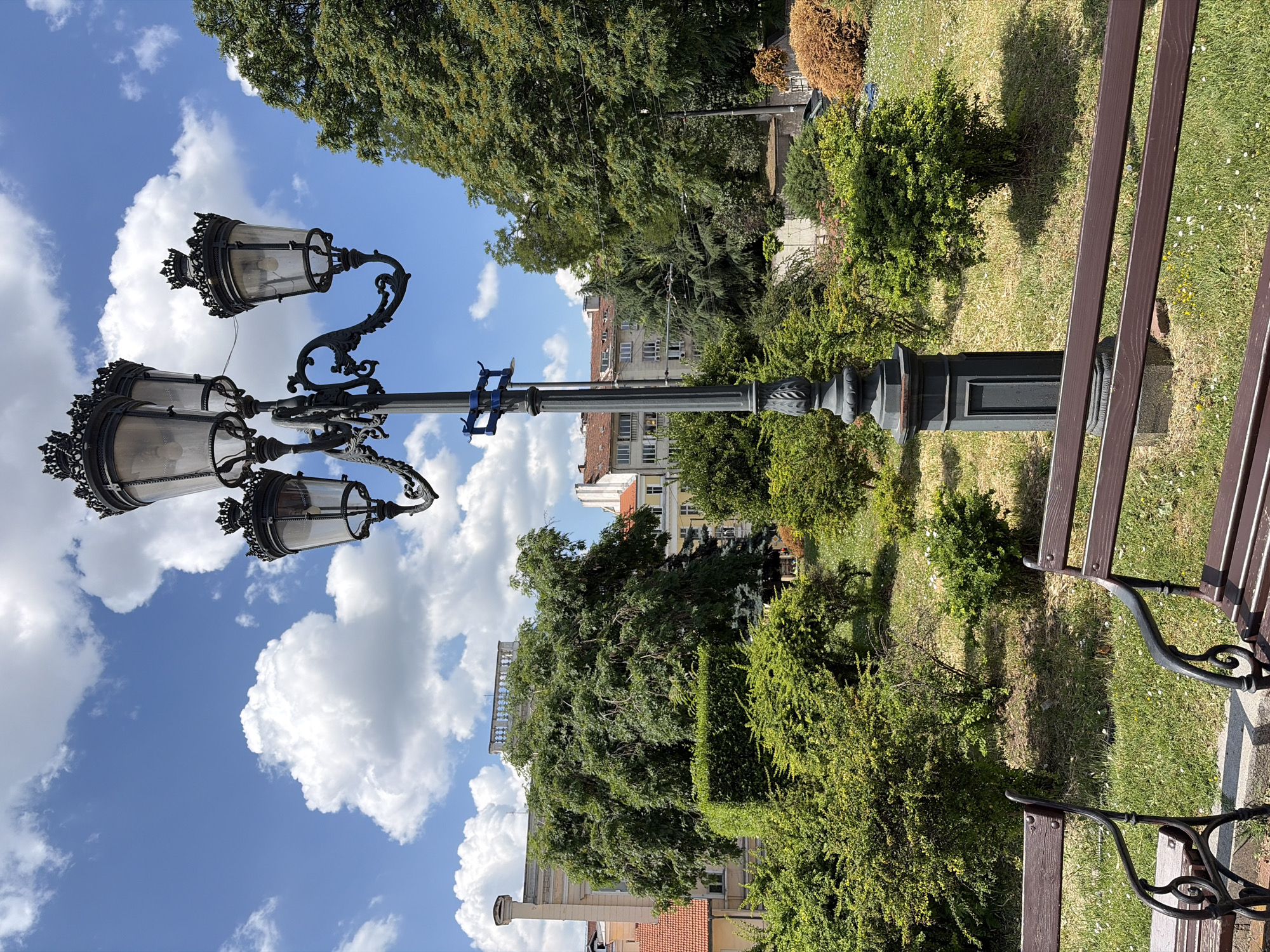 A tall, ornate lamppost with three lamps stands in a park on a sunny day. The lamppost is dark gray and made of metal, with decorative scrollwork. The lamps are clear glass with black decorative tops. Behind the lamppost, there are several benches, lush green bushes, and trees. In the background, there are buildings with red roofs and balconies. The sky is blue with fluffy white clouds. The scene appears to be in a city park, possibly in Sofia, Bulgaria, given the architectural style of the buildings and the lamppost.