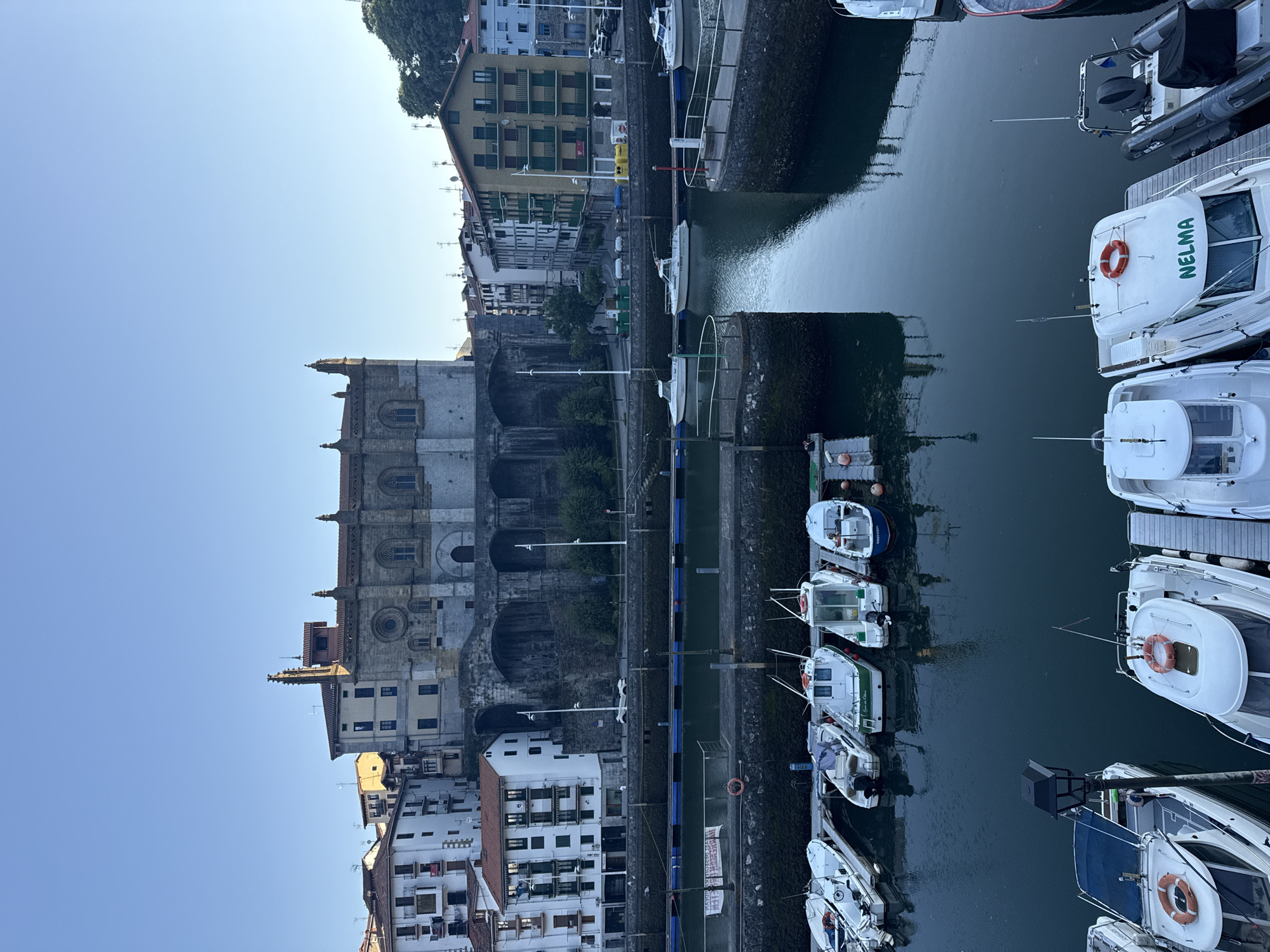 The image captures a harbor scene in Ondarroa, Spain, with a clear blue sky overhead. A prominent stone building, likely a church or cathedral with Gothic architectural elements, stands on a raised bank overlooking the water. Several multi-story buildings with residential facades line the waterfront. In the foreground, numerous boats are docked in the harbor. These include smaller motorboats, some with white hulls and green trim, and a larger inflatable boat with a dark tube. The water is calm, reflecting the surrounding structures. The scene is one of a peaceful morning in a coastal town.