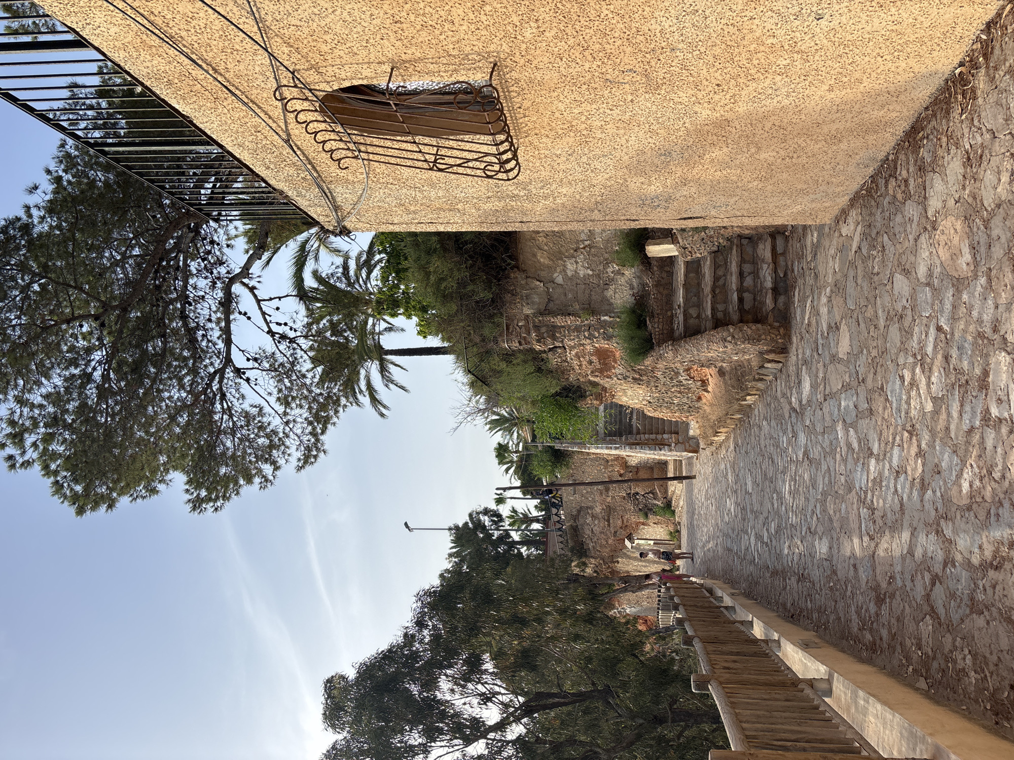 A person is walking down a stone path lined with a wooden fence on the left. Two people stand in the distance on the path. The path is flanked by a rock wall and vegetation on the right, leading to a set of stone stairs. A building with a textured yellow facade and a barred window is visible on the right side of the image. Palm trees and other foliage are present on the hillside above the path. The sky is a pale blue with wispy clouds.
