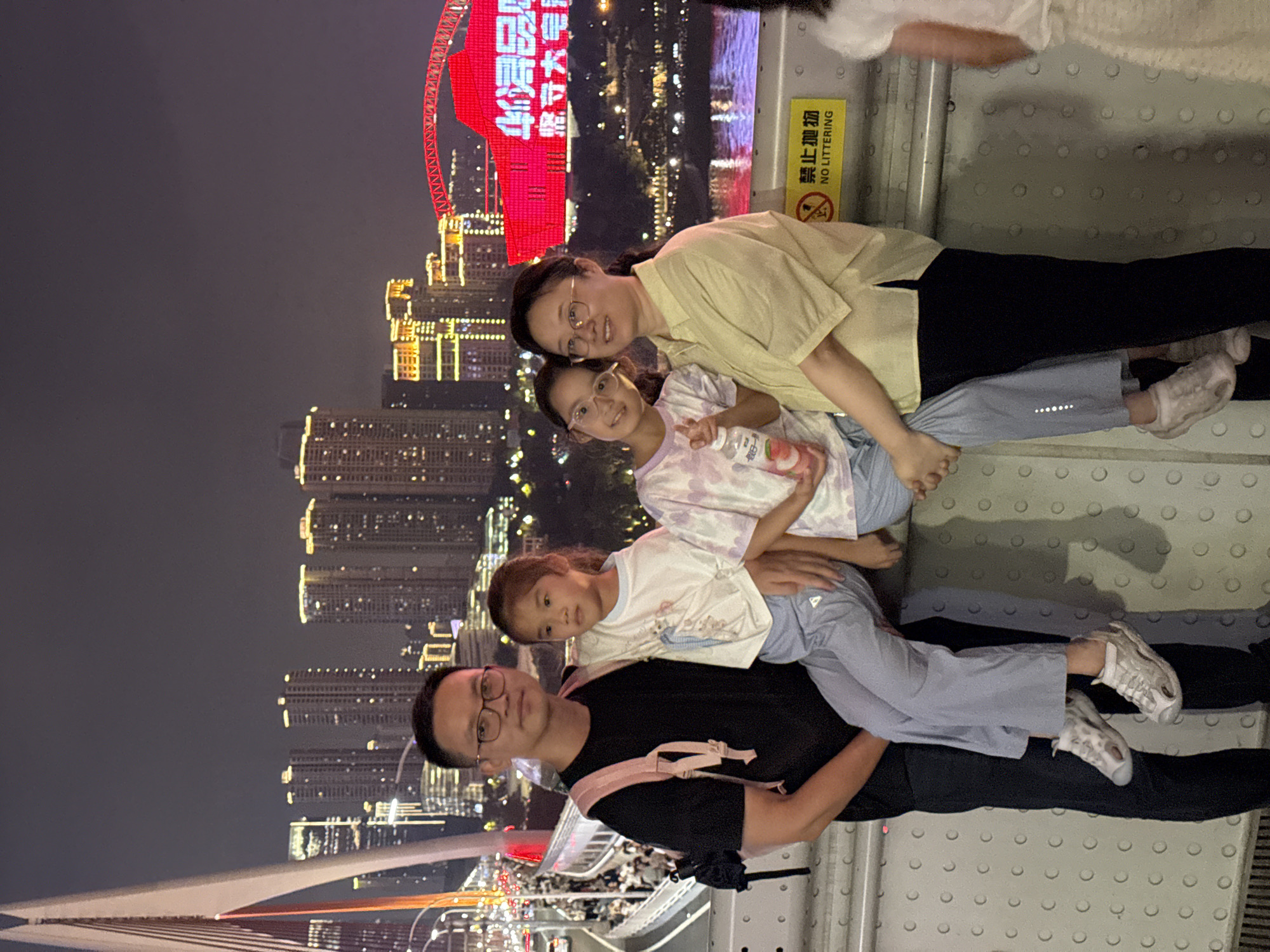 An outdoor nighttime photograph taken at the Hongyadong scenic area in Yuzhong District, China, depicts a family of four standing on a bridge or walkway. On the left, a man wearing glasses, a black t-shirt, and dark pants, with a pink backpack on his shoulders, holds a young girl on his left arm. The girl, wearing a white t-shirt and light blue pants, looks off to the side. Next to them, another young girl, also wearing glasses, a tie-dye t-shirt, and light blue pants, is seated. She holds a bottle of peach-flavored drink. On the right, a woman wearing a light yellow short-sleeved shirt and black pants stands with her arm around the seated girl. Behind the family, a cityscape is visible, illuminated by numerous building lights and a large red structure. The background also shows a body of water. A yellow sign with Chinese characters and the words "NO LITTERING" is visible on the right.
