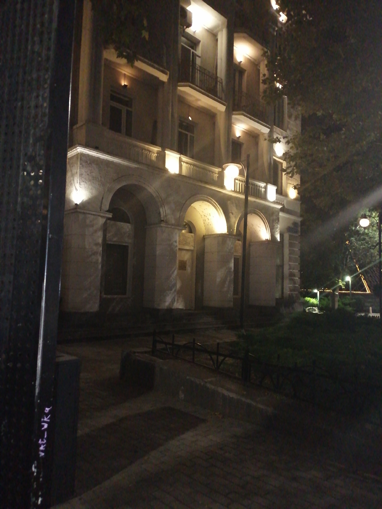 The image is a night-time photograph of a building exterior in Rustavi, Georgia. The building appears to be multi-story with several arched entrances on the ground floor and balconies on the upper floors. The facade is illuminated by artificial lights, highlighting architectural details such as columns and decorative railings. Trees with dense foliage are visible to the right of the building, and a dark metal fence is in the foreground to the left. The ground in front of the building is paved, and there is a grassy area with some lighting and possibly a bench in the background. The overall atmosphere is dark and somewhat moody due to the night setting and artificial lighting.
