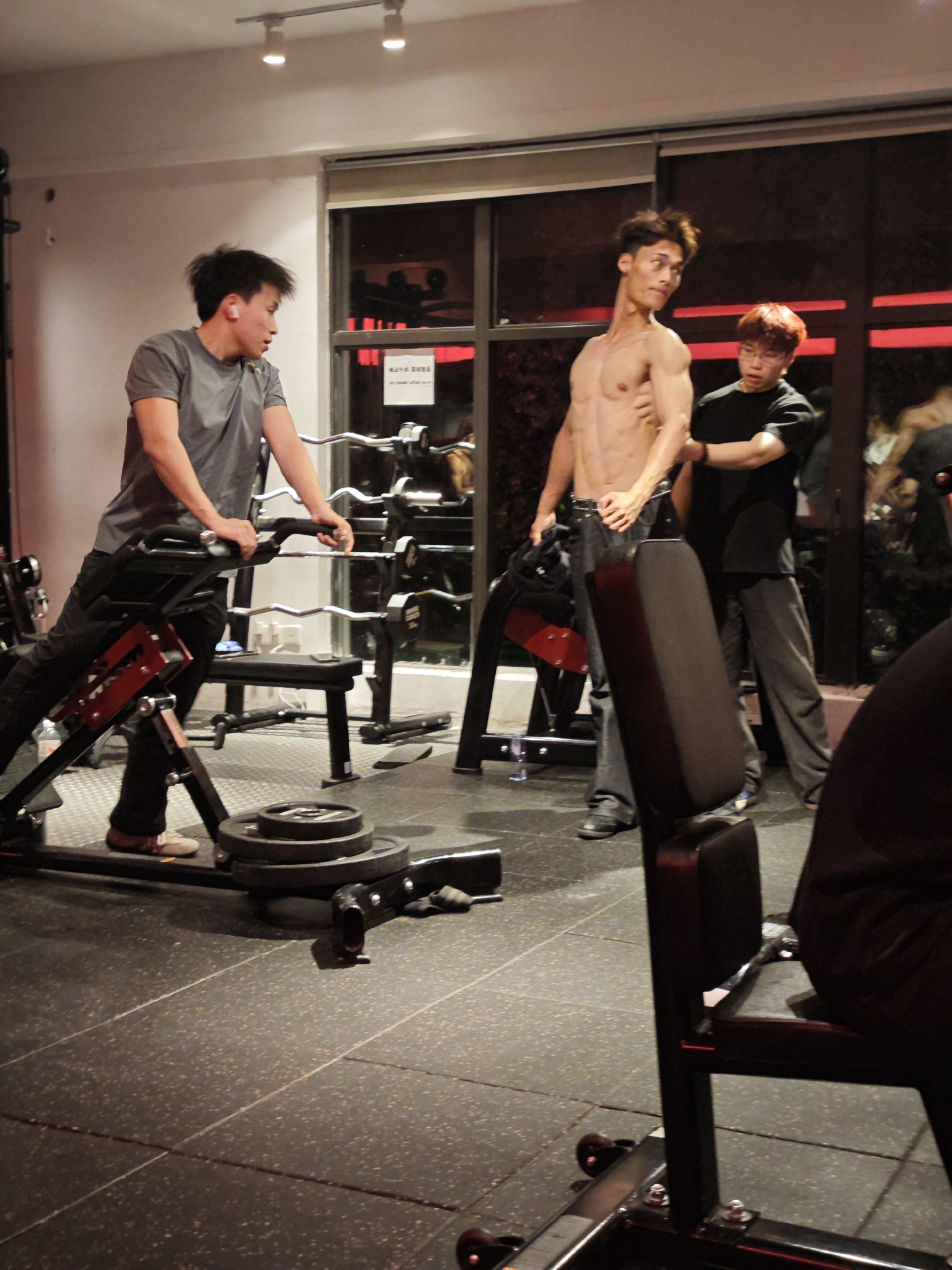 A group of young Asian men are in a gym, possibly located in the Chongqing High-tech Zone, China. The lighting in the gym is dim, with red accents from the background lights. The floor is a dark, textured material.
In the foreground, a man with dark hair is using a weight machine. He is wearing a gray t-shirt and black pants, and is leaning forward, holding onto the machine's handles. A stack of weight plates is visible on the machine.
To the right, a shirtless man with a lean, muscular physique is standing and posing. He has dark hair and is looking towards the camera. Another man, wearing a black t-shirt and gray pants, stands next to him, with his hand on the shirtless man's arm, possibly assisting or observing.
In the background, there are several weight racks with barbells and weights, suggesting a well-equipped gym. A window or glass partition in the background shows reflections and the red lighting. There are also signs visible on the wall, but their content is not legible. The overall impression is a casual gym session among friends.