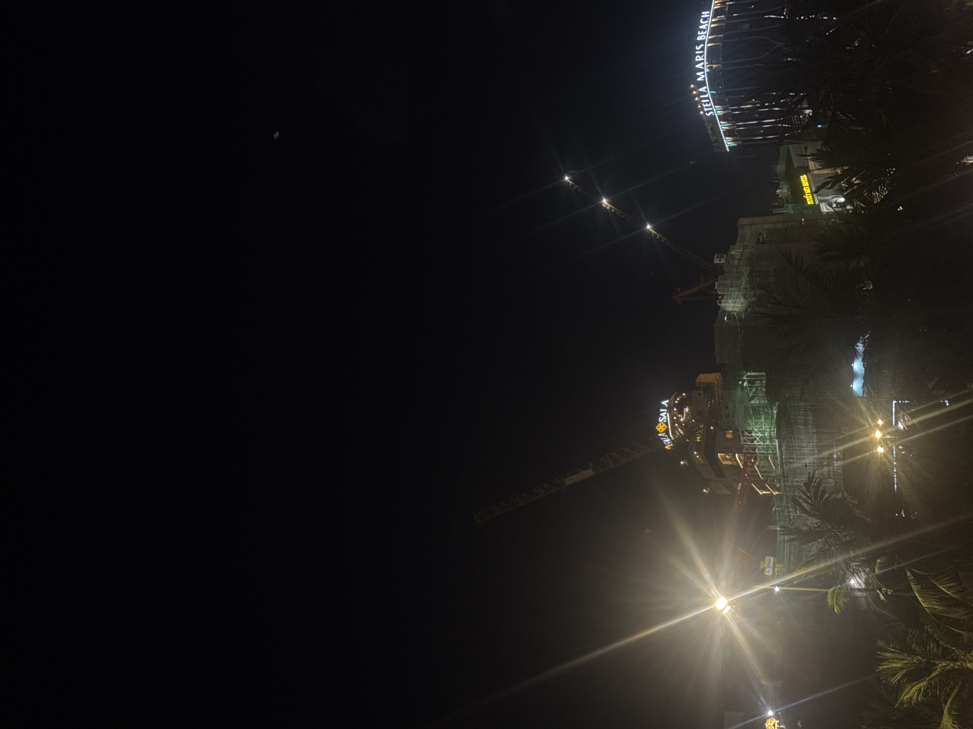 The image shows a nighttime scene in Đà Nẵng, Vietnam, with a focus on ongoing construction. In the lower-right quadrant, palm trees are visible against a night sky. Prominently featured are construction sites illuminated by bright lights. Cranes are visible, indicating ongoing work on buildings, one appears to be named "SALA". In the upper right of the image, the illuminated signage for "STELLA MARIS BEACH" is visible. Additional hotel signage is evident.