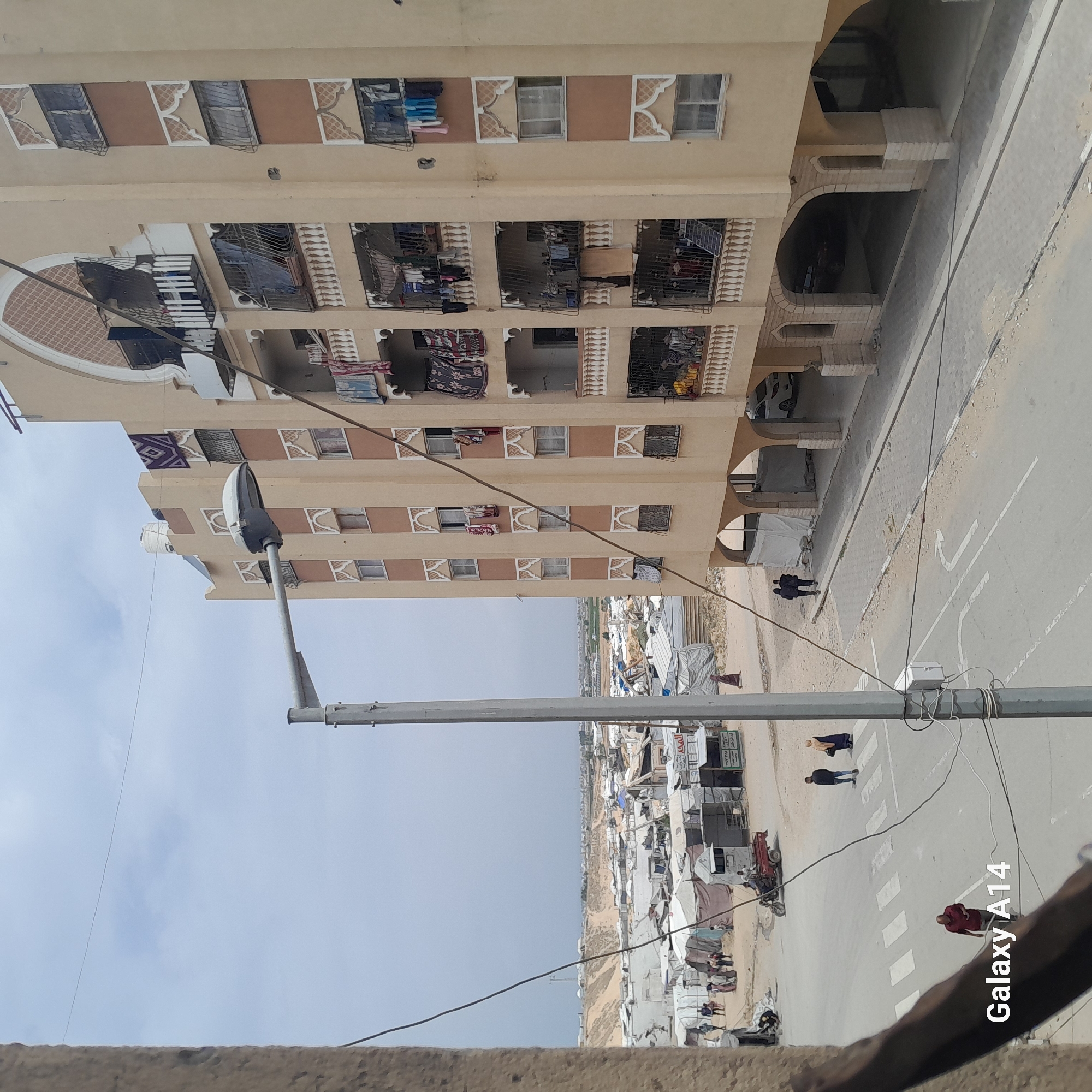 The image shows a multi-story building with light-colored facade and several balconies, some with items hanging on them. Next to the building is a road with visible lane markings and a pedestrian crossing. Several people are walking on the road and sidewalk. In the background, there is a large area covered with what appears to be makeshift shelters or tents, suggesting a camp or unplanned settlement. The weather is overcast. The image has "Galaxy A14" printed at the bottom. The city of Unknown is unknown.
