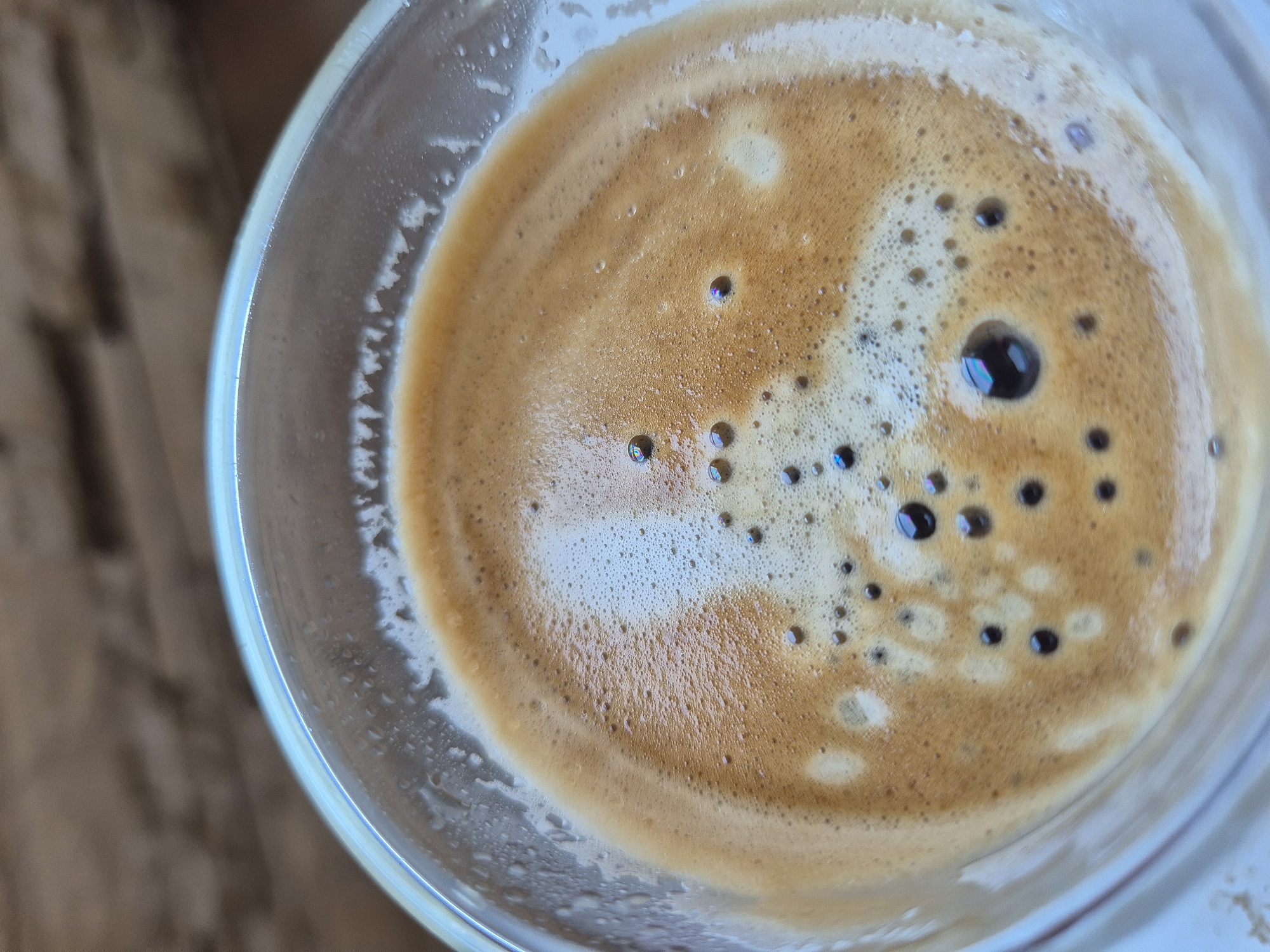 An overhead shot captures a clear glass cup filled with coffee, likely an espresso, showcasing its frothy crema. The surface of the coffee is a vibrant mix of light brown and golden-brown tones, forming intricate patterns and textures. Numerous small bubbles are scattered across the crema, while a few larger bubbles appear darker due to reflections, some even exhibiting subtle iridescent hues.
The rim of the clear glass cup is prominently visible, and its edge displays a slight blue tint from light refraction. Along the interior of the glass, just above the coffee line, there are visible traces of dried foam or coffee residue. The background, largely out of focus, reveals warm, blurred brown tones, suggesting a wooden surface such as a table or countertop. The lighting appears bright, highlighting the details of the coffee and the transparency of the cup. There are no people visible, nor any discernible text. The image portrays a tranquil moment, perhaps before the coffee is consumed.