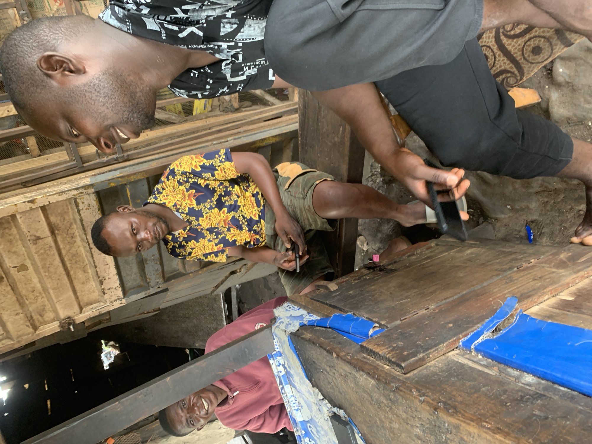 In Shagamu, Nigeria, three men are gathered in what appears to be an open-air market stall. The man on the left, wearing a maroon hoodie, is smiling and looking at the camera. The man in the center, dressed in a floral shirt and shorts, is holding a mobile phone and looking down at it. The man on the right, wearing a black and white patterned shirt and black pants, is also looking to his left and smiling. The environment is rustic, with wooden structures and a dirt floor. Blue plastic sheeting is visible on a wooden surface in the foreground, and some debris is scattered around.