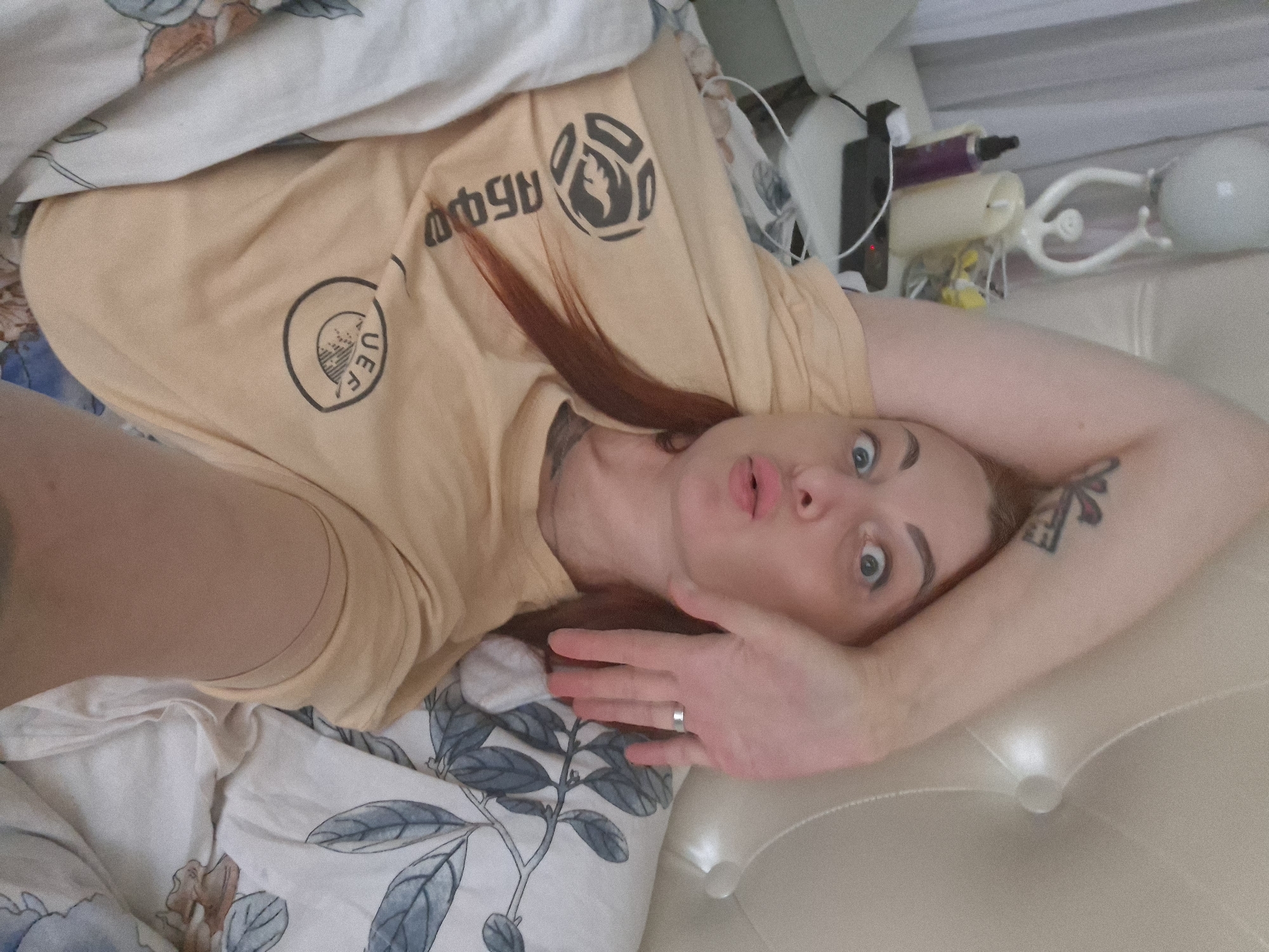 A woman with auburn hair is lying in bed, looking at the camera with a surprised expression and puckered lips. She is wearing a light orange t-shirt with a logo that appears to be the UEFA Europa League logo. Her left arm is raised above her head, and her right hand is resting on her face. The bed is covered with white and floral patterned sheets. In the background, on a nightstand, are a lamp, a candle, and some other objects. The overall impression is that the photo was taken in a bedroom, likely in a home environment, and given the Myadzyel, Belarus context, it is a residential bedroom in that city.