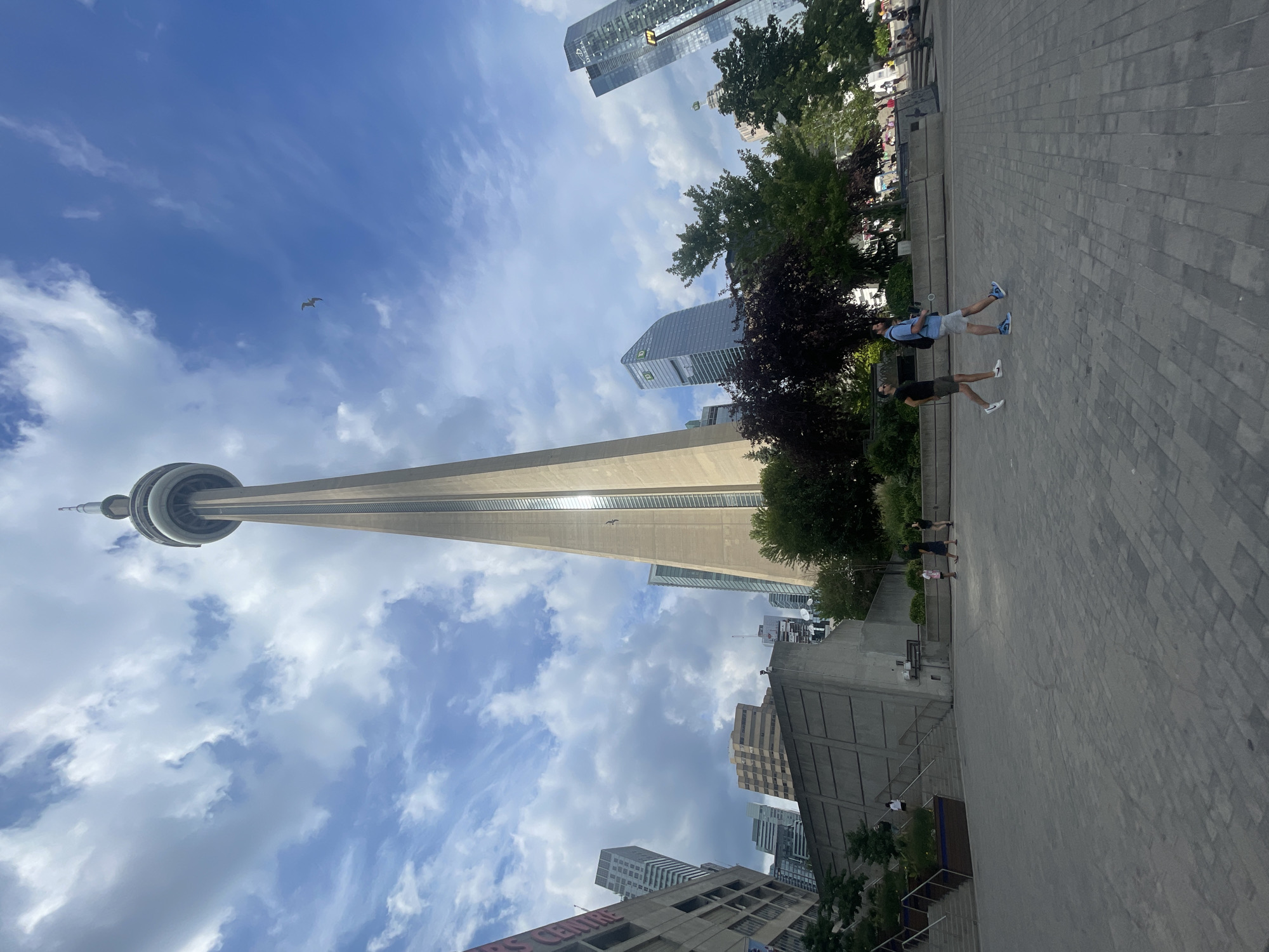 The image depicts the CN Tower in Toronto, Canada, on a partially cloudy day. The CN Tower is the dominant structure, towering over the surrounding cityscape. In the foreground, a paved plaza with a brick-like pattern is visible. Two men are walking across the plaza, one in a black t-shirt and khaki shorts, the other in a blue t-shirt and grey shorts. To the left of the tower, a concrete structure with stairs and planters is present. Several other modern skyscrapers, including the TD Bank buildings, are visible in the background, interspersed with trees. A bird is seen flying in the sky near the top of the CN Tower.