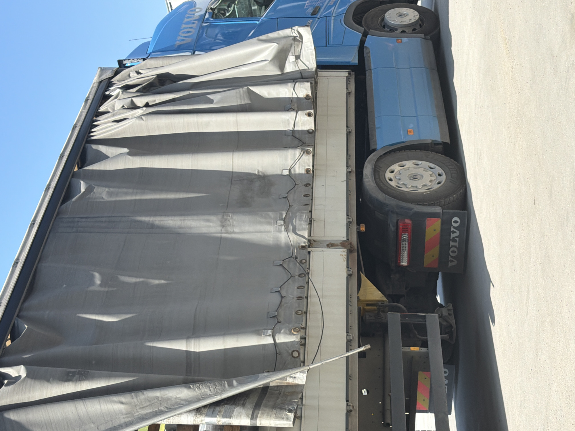 A blue Volvo truck is parked on a paved surface, possibly a road or lot, under a clear sky. The truck's trailer is covered with a large, grey tarp, which is partially pulled back, revealing the metallic side of the trailer. The tarp is wrinkled and creased, suggesting it has been used. The truck's cab is visible in the background, also blue, with "VOLVO" written in large white letters on its side. The wheels of the truck are prominent, one of them showing a silver hubcap. The rear of the truck has red and yellow reflective markings and "VOLVO" lettering. The overall scene appears to be outdoors during the daytime, with natural light casting shadows on the ground and the side of the truck. The setting appears to be a relatively unpopulated area, given the focus on the truck and the simple, paved ground. There are no people visible in the image.