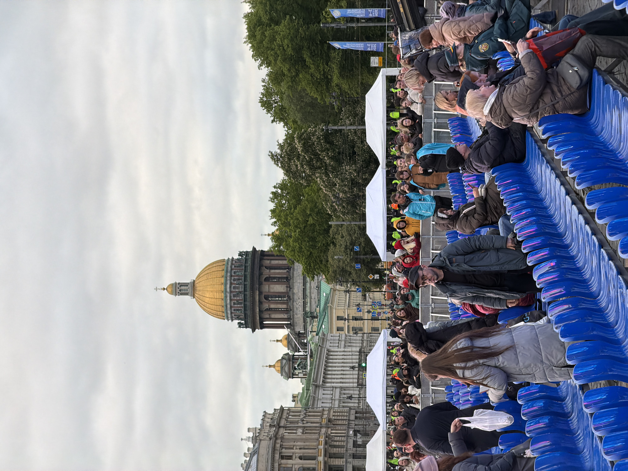 From the perspective of someone in stadium seating, the image shows a crowd of spectators looking towards Saint Isaac's Cathedral in Saint Petersburg, Russia. The sky is overcast. White tents are interspersed among the crowd and trees. The blue plastic stadium seats appear to be mostly occupied by people in neutral-colored clothing. Many spectators are looking at their phones.
