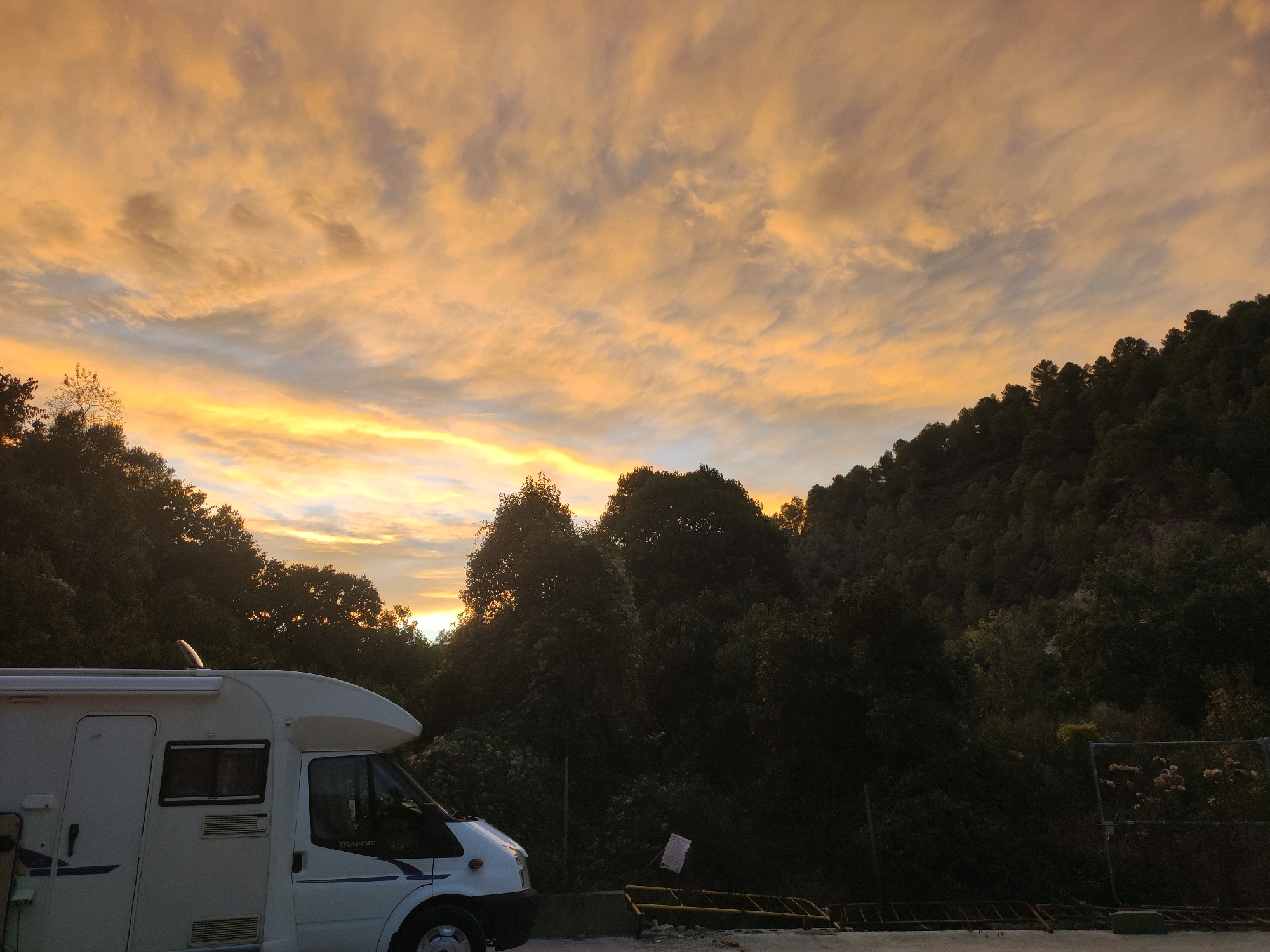 An RV is parked in front of a densely wooded hillside under a dramatic, cloudy sky with hues of orange, yellow, and gray. The sky is illuminated by what appears to be the setting or rising sun, with light breaking through the clouds and casting an atmospheric glow. The RV is white and appears to be a camper van or motorhome, positioned on a paved area. The hillside is covered in dark green trees, creating a silhouette against the brightly lit sky. In the foreground, some yellow barriers and debris are visible, suggesting a parking area or roadside location. The overall scene depicts a serene, natural setting with a human-made element, possibly a campsite or a stopover point. The image implies a location in a rural or semi-rural area, with the presence of natural landscape. The specific location in Suera, Spain is not discernible from the visual information alone.