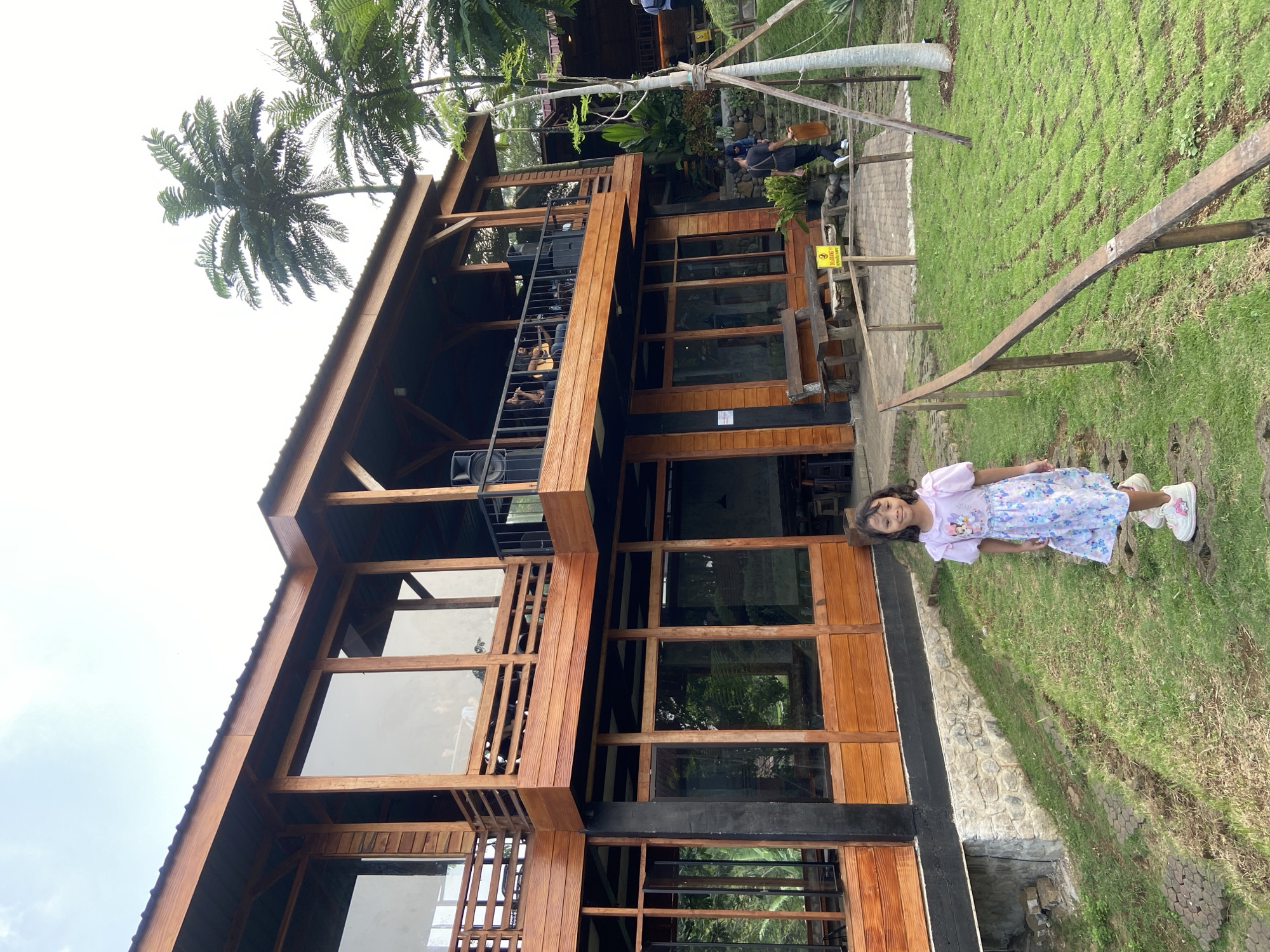 A girl stands in a grassy area next to a three-story wooden building with large windows. The building has a black roof, brown window frames, and a stone foundation. A palm tree is visible to the left of the building. The scene is located in Tamiajeng, Indonesia.