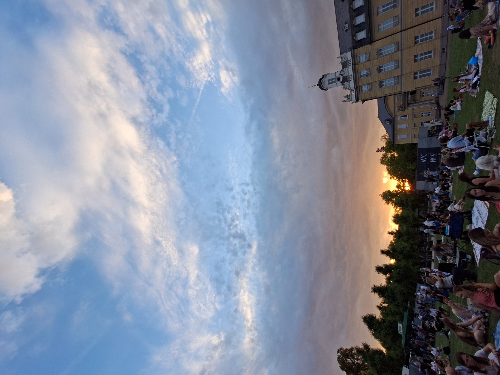 The image shows a wide shot of an outdoor gathering at sunset in Karlsruhe, Germany, likely at the Karlsruhe Palace. A large crowd of people are seated on a grassy lawn, facing away from the camera, some in lawn chairs and others on blankets. In the background, a large yellow building with a distinct tower and numerous windows dominates the right side of the frame. Trees line the middle ground, and a faint outline of a stage or screen can be seen on the left. The sky above is a mix of blue, with streaks of white and grey clouds, and the setting sun casts a warm orange glow towards the horizon. The overall atmosphere suggests an evening event, possibly an outdoor concert or movie screening.