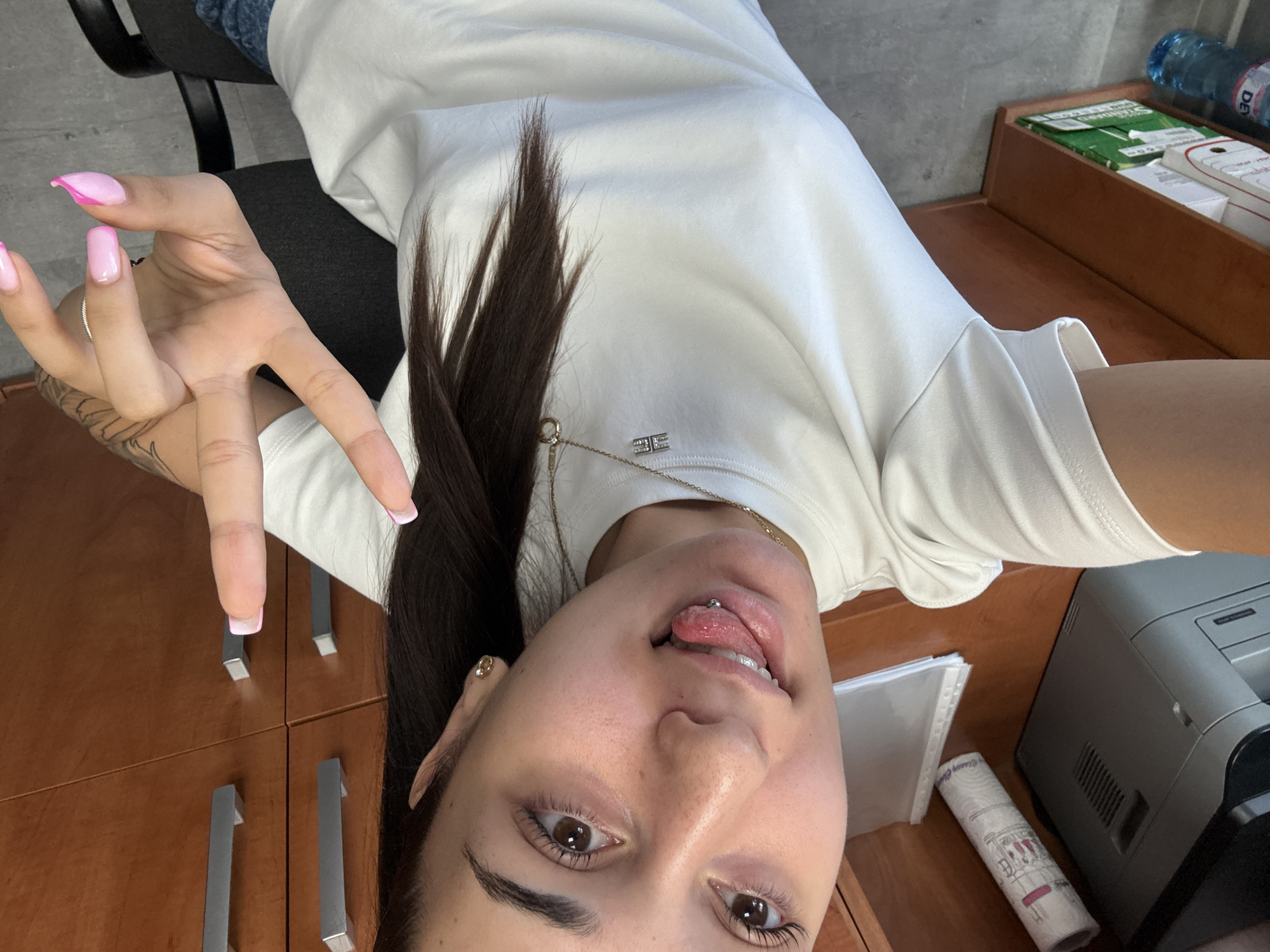 A young woman with dark hair and a tongue piercing is taking a selfie in an office environment. She is wearing a white t-shirt and a necklace with a pendant. Her right hand is raised in a peace sign, showing off her pink nail polish. In the background, there are wooden cabinets and office equipment, including a printer and some folders. A bottle of water is visible on the floor. The scene suggests a casual moment in a workplace. The location is identified as Dibich, Bulgaria.