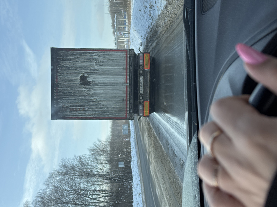 The image presents a driver's perspective from inside a vehicle on a winter road in L'vovskiy, Russia. In the bottom-left foreground, a light-skinned hand, exhibiting two rings on the ring finger and pink-painted fingernails, grips a black steering wheel. Directly ahead, a dark semi-trailer occupies the central frame, viewed from the rear. The trailer's surface is heavily streaked with a pale, possibly frozen or dried, residue. A Russian license plate, "РВК 4648 77," is affixed to the lower rear of the trailer. The asphalt road surface appears wet and reflective, with visible tire tracks, indicating moisture or icy conditions. Snow covers the ground along the roadside verges. To the left, a line of bare deciduous trees and some evergreens are visible, partially covered in snow. The sky is predominantly clear blue with sparse white clouds. In the distant background on the right, an elevated white structure, consistent with a gas station canopy or a toll booth, is discernible.