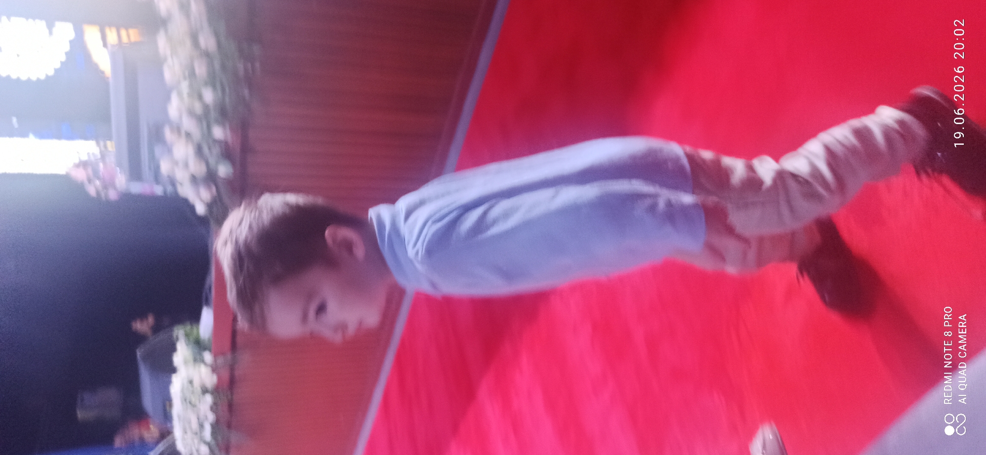 An out-of-focus, full-body side view of a young Caucasian boy walking on a red carpet. He is wearing a light blue collared shirt and tan pants. His dark hair is short and styled. The boy is in motion, appearing to walk to the left of the frame. Behind him, a dark wooden wall is visible, with some blurred floral arrangements and stage lighting in the background. The bottom right corner of the image contains a timestamp: "19.06.2026 20:02".