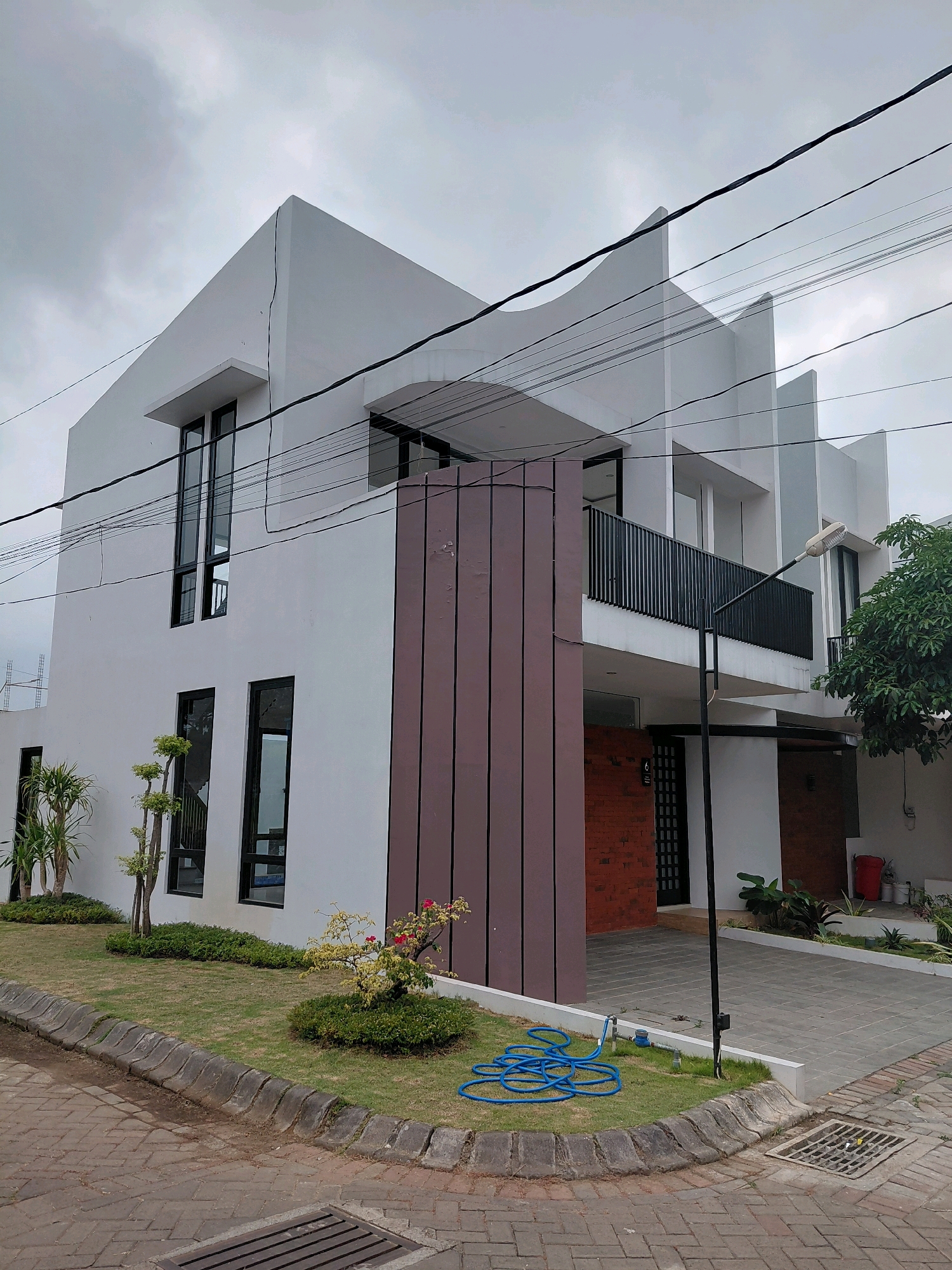 A modern, two-story white house with sharp geometric angles and black-framed windows is situated in a suburban area of Batu, Indonesia. The house features a balcony with a black metal railing on the second floor. A prominent brown vertical panel is affixed to the exterior wall. The grounds include a manicured lawn with small ornamental trees and shrubs, a paved driveway, and a brick-paved sidewalk. A blue garden hose is coiled on the grass near the sidewalk. Overhead, power lines traverse the cloudy sky. The overall impression is of a new construction in a residential neighborhood under an overcast sky.