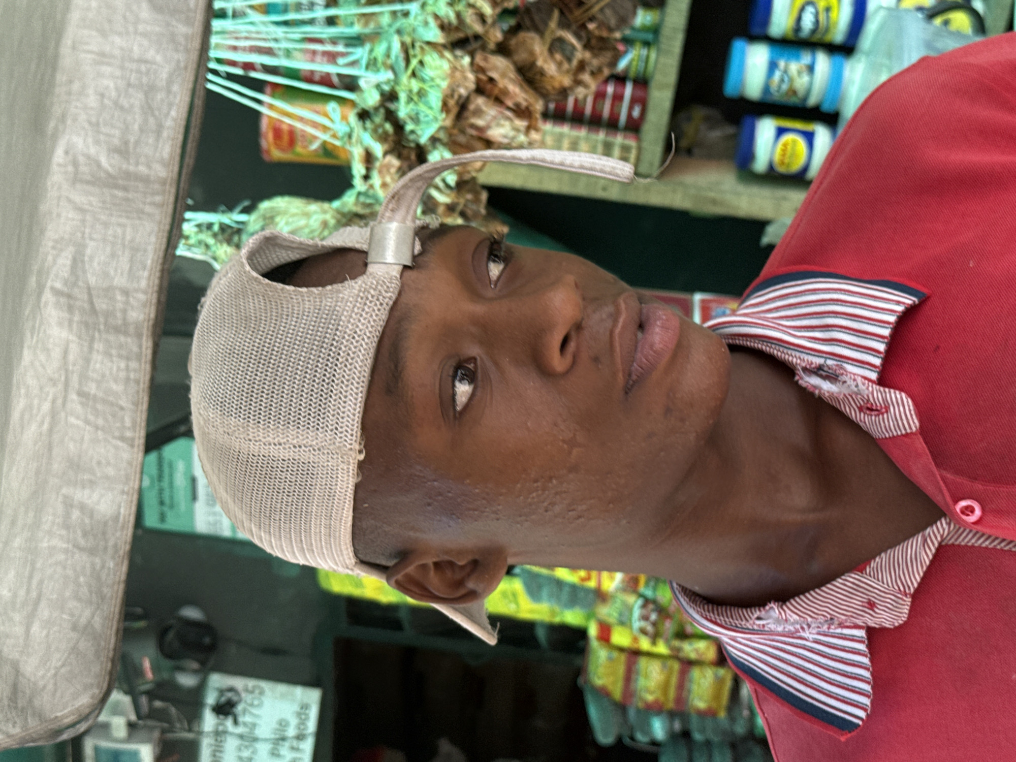 A close-up, medium shot captures a dark-skinned man wearing a tan, mesh baseball cap with an unusually long and curled adjuster strap. He is also wearing a red collared shirt with a white and red striped trim. The man is looking slightly upward with a neutral expression. The background is cluttered with various objects and food items, some hanging on display and others stacked on shelves. A sticker with the words "niaps 4304765" and "P Nile Foods" is visible. The location is likely a small shop or market stall in Ado, Nigeria.