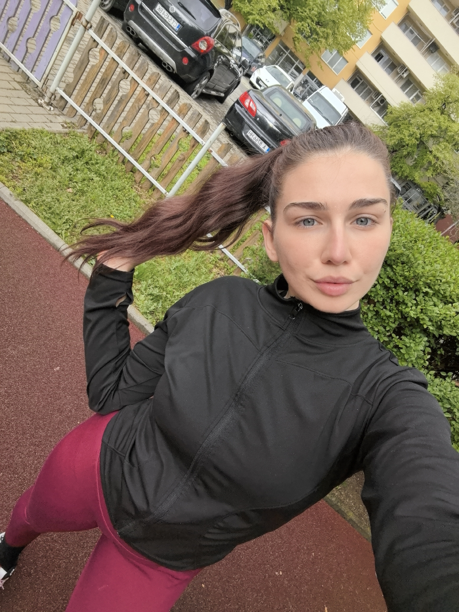 A woman is taking a selfie outdoors. She has light skin, blue eyes, and her brown hair is in a high ponytail. She is wearing a black long-sleeved athletic jacket and burgundy leggings. The background includes a wooden fence, green bushes, parked cars, and a multi-story building. The ground is a reddish-brown color. The photo appears to have been taken on a walkway next to a street. The location is likely in Stara Zagora, Bulgaria, given the context.