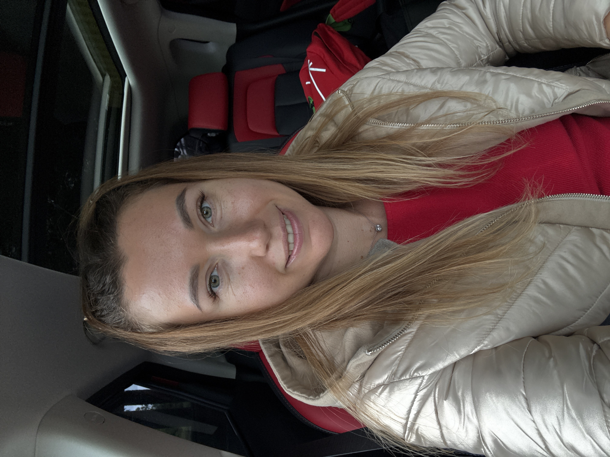 A woman with blonde hair and fair skin is sitting in a car. She is wearing a red sweater and a beige puffer jacket. She is smiling. The car has red and black seats. There is a red backpack in the background. The location is unknown.
