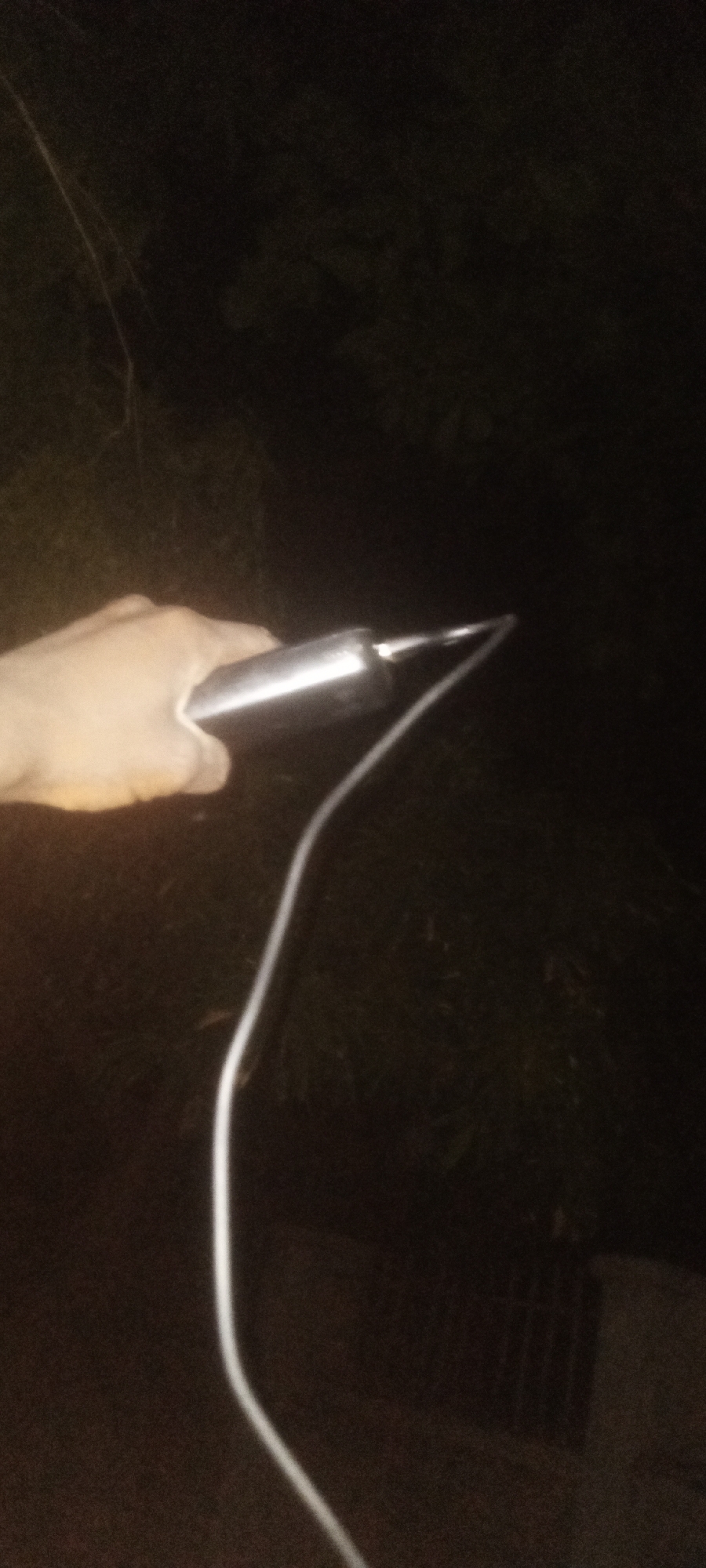 A hand holds a cell phone connected to a white charging cable in a dark outdoor environment. The setting appears to be Sabon Tasha, Nigeria, judging by the location tag (although the environment itself can not be determined by the image). Vegetation is faintly visible in the background.