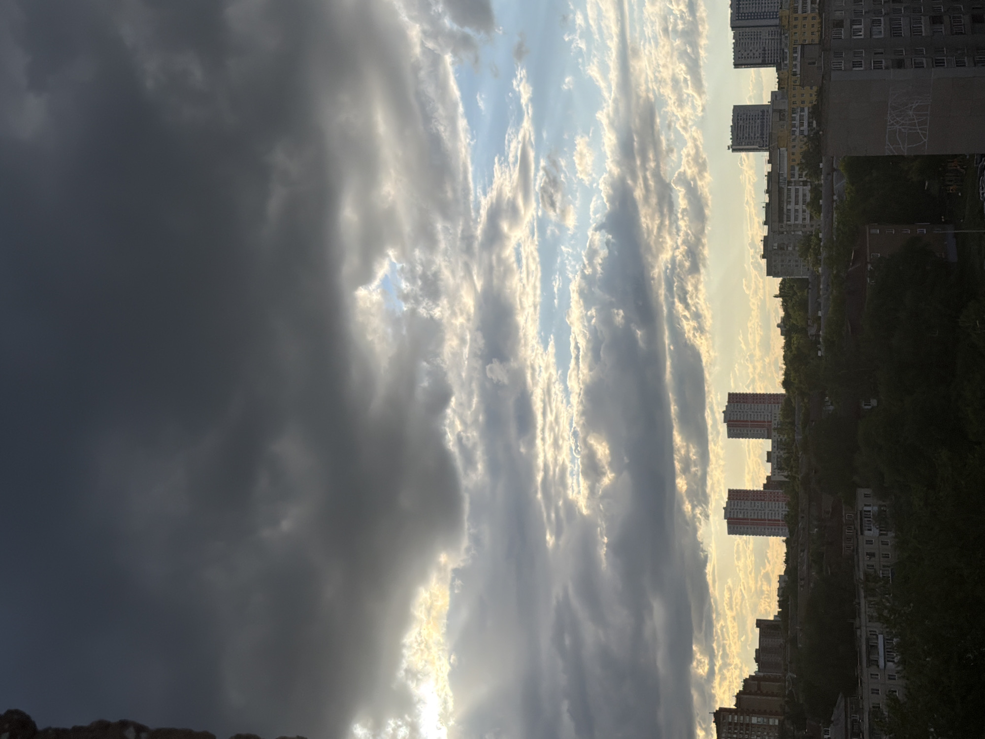 The image shows a cityscape under a sky transitioning from a dark, overcast area to a brighter, sunlit zone with striated clouds. On the right side, tall residential buildings of varying heights line the horizon, partially obscured by trees. The buildings appear to be typical urban apartment blocks. The left side of the sky is dominated by dark storm clouds. The environment appears to be a city or urban area. There are no visible people or interactions in the image. The buildings are located in an urban area of unknown city.