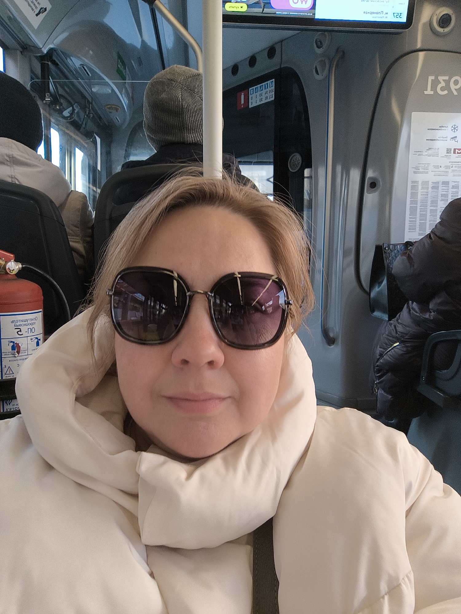 This media file depicts a woman taking a selfie inside a public transport vehicle, likely a bus, in Khimki, Russia. The main subject, positioned in the foreground, is looking directly at the camera, wearing large dark sunglasses that obscure her eyes and a light-colored (cream or off-white) puffer jacket. Her light blonde hair is visible around her face.
Behind her, the interior of the bus shows two other passengers seated. One passenger is seen from behind, wearing a grey knitted beanie, while another is partially visible further back. The bus interior features standard grey plastic seating, vertical metal poles for standing, and clear partitions.
Notable objects include a red fire extinguisher mounted to the left of the main subject, clearly labeled with the Cyrillic text "ОГНЕТУШИТЕЛЬ" (Fire extinguisher) and "В-ОП 2-ПО", along with illustrative safety instructions. An overhead digital display screen shows the number "321", likely a route number, and the Cyrillic word "вперед" (forward), confirming the Russian context. Other visible text on the display includes "KAUNLP" and "SOUKY". Various informational signs and advertisements are affixed to the bus walls, displaying numbers such as "102 34 30 10" and small icons, including a snowflake and a mail envelope. The lighting suggests it is daytime. The woman's visible mouth indicates a neutral or slightly pleasant expression.
