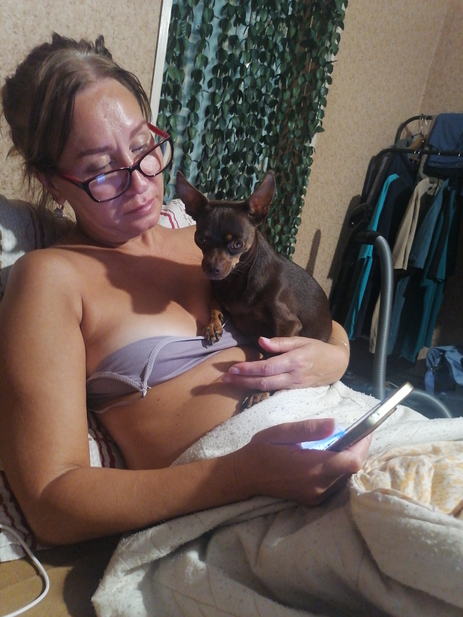 A woman with brown hair pulled back is wearing glasses and a light purple strapless bra. She is holding a small brown dog on her chest. She is looking at a smartphone in her right hand, which is glowing with blue light. The background shows a wall with a green vine-like decoration, a clothes rack with several garments, and a vacuum cleaner hose. The scene appears to be indoors, likely in a bedroom or living room. The woman is lying down, covered by a blanket.