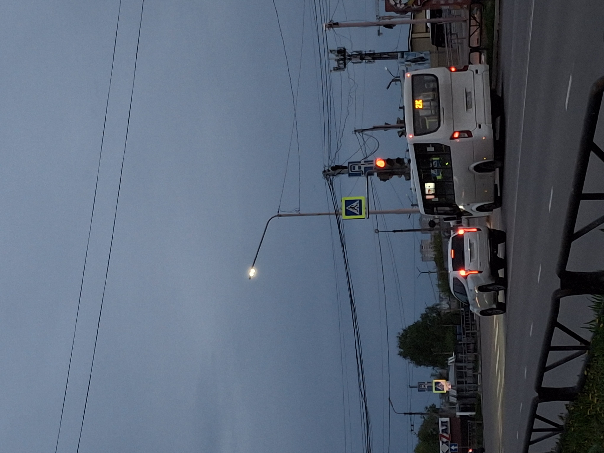 The image shows a street scene in the city of Chaikovsky, Russia. A white bus with the number "2K" displayed on its rear is stopped at a red traffic light. A white car is behind the bus, also stopped at the light. A pedestrian crossing sign is visible next to the traffic light. Streetlights and power lines are present in the background, and some trees and greenery are visible along the side of the road. The sky is overcast. A metal guardrail is on the right side of the image.