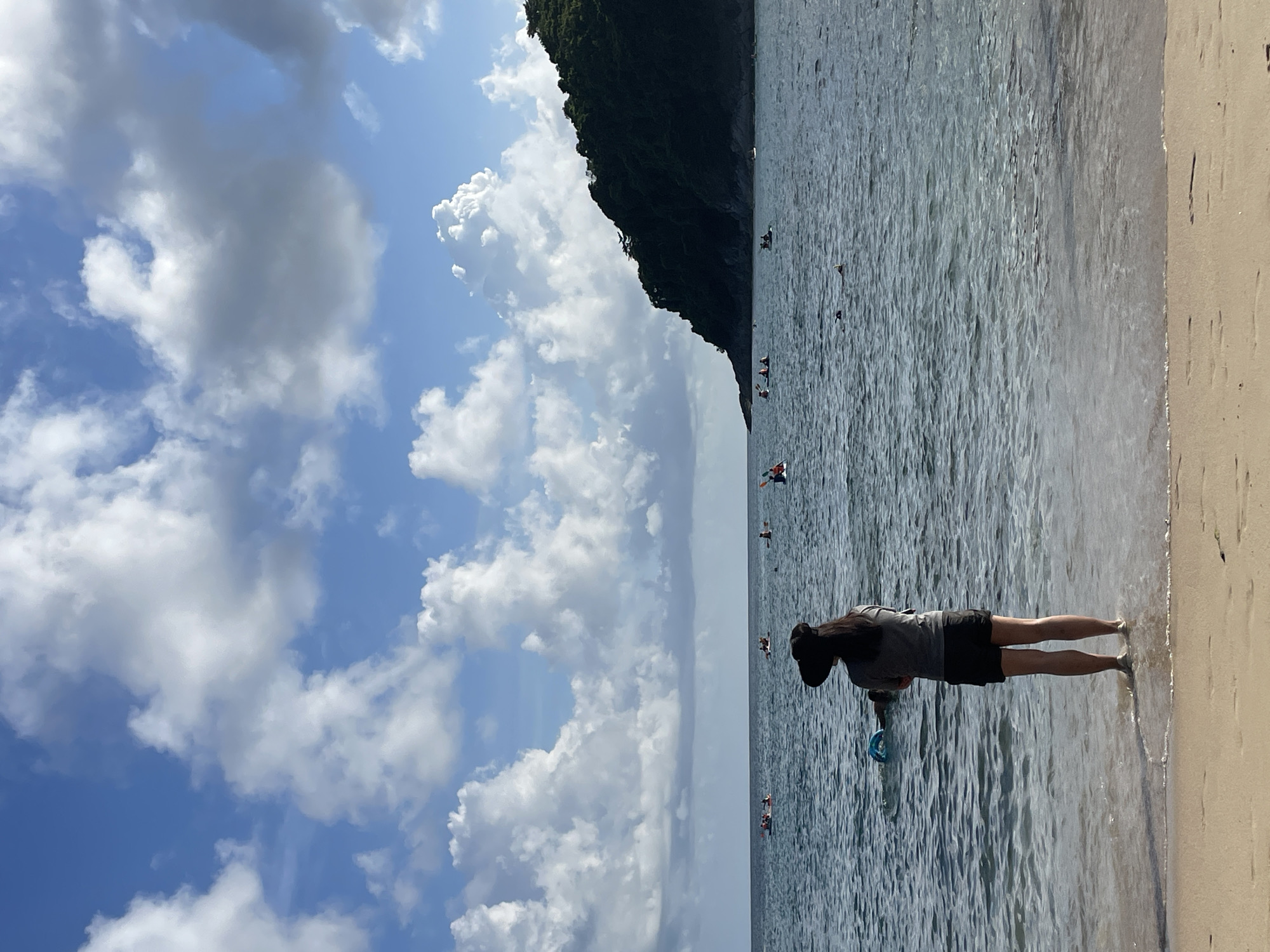 The image depicts a serene beach scene in Rawai, Thailand, under a sky filled with dynamic cumulus clouds against a vibrant blue backdrop. In the foreground, a person stands at the water's edge, facing the sea. They are wearing a wide-brimmed hat, a grey t-shirt, and dark shorts. Their feet are submerged in the shallow, clear water, and they are holding a blue circular object.
The sea stretches out before them, calm with gentle waves lapping at the shore. Further out in the water, several individuals are engaged in water activities, with some seen in kayaks and others seemingly swimming or snorkeling. A lush, green, tree-covered hill forms the backdrop on the right side of the image, with its dark silhouette contrasting against the bright sky and sea. The beach itself is composed of light-colored sand. The overall impression is one of a peaceful day at the coast, with various people enjoying the ocean and the natural beauty of the location.
