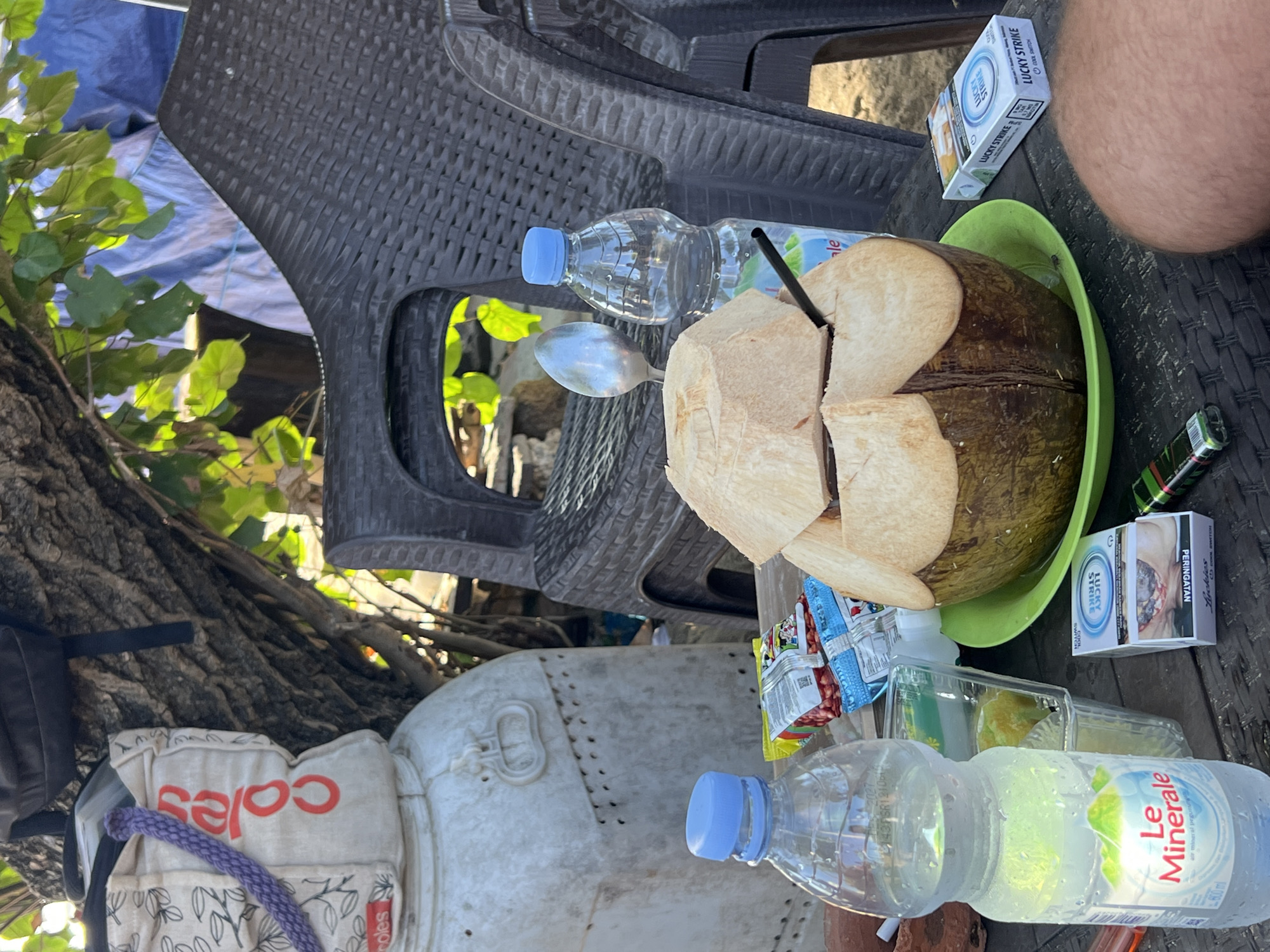 A close-up, eye-level shot shows a table setting with refreshments and smoking accessories under the dappled shade of a tree, likely in Kuta, Indonesia. In the foreground, a large, freshly opened coconut sits on a green plate, its top half removed to reveal the white flesh. A black straw is inserted into the coconut. To the left of the coconut, a bottle of "Le Minerale" water is prominently displayed, its label featuring mountains and the brand name in red and white. Adjacent to the water bottle are several small, foil-wrapped snack packets. To the right of the coconut, a pack of "Lucky Strike" cigarettes, featuring Indonesian warning labels, rests on the table. A lighter is also visible near the cigarette pack. In the background, a dark plastic chair is partially visible, and beyond that, a large white container with metal accents and a faded "Coles" logo on a canvas bag is situated. The overall impression is a relaxed, outdoor setting, possibly a beachside cafe or a private patio, with a focus on tropical beverages and items associated with leisure.