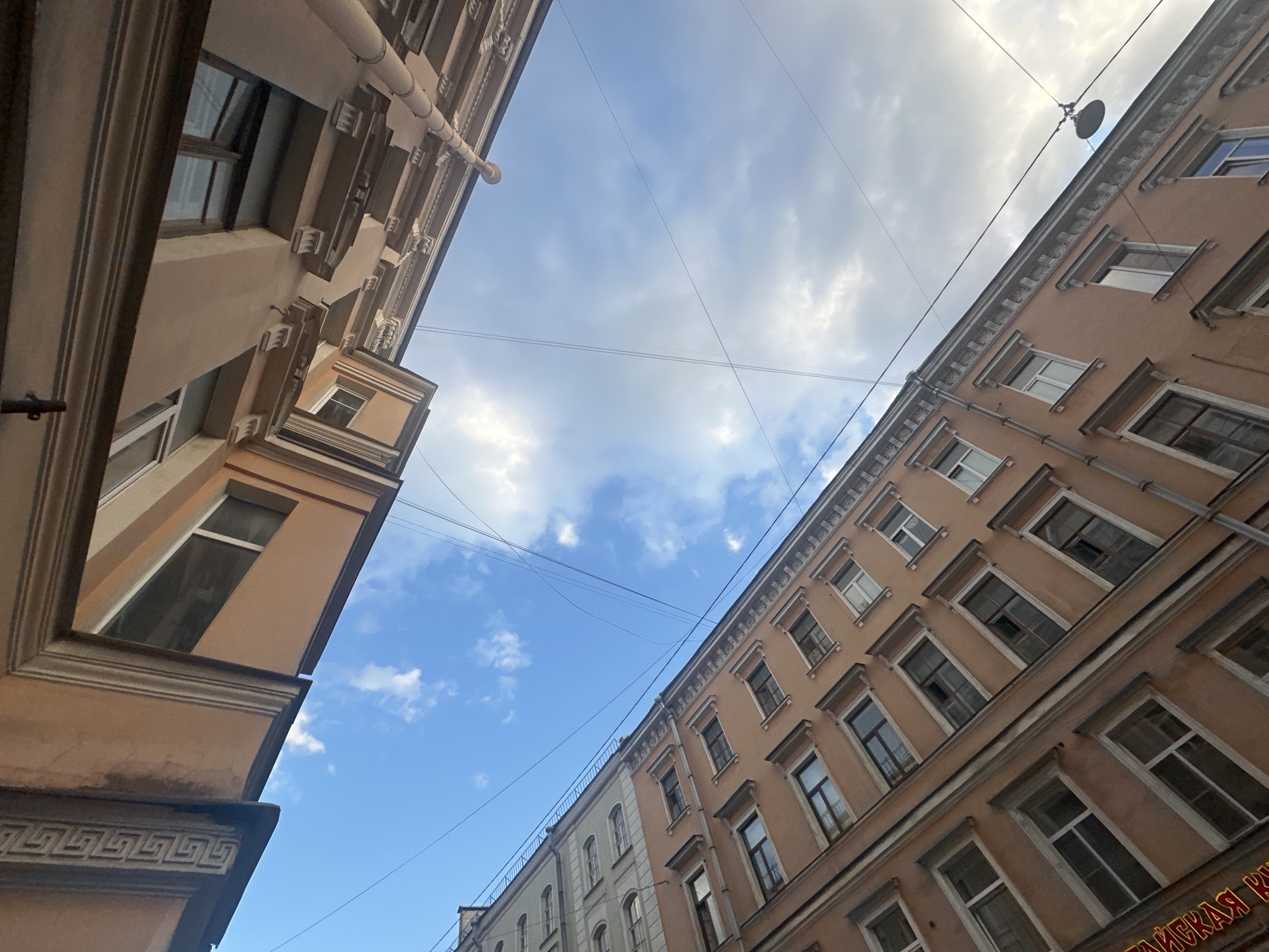 The image shows a low-angle view looking up at two buildings in an urban environment. The buildings are tall and multi-storied, with facades made of pale orange and light gray stucco. Both buildings feature numerous windows, many with white frames, set within a decorative architectural style that includes cornices and molding. The sky is visible between and above the buildings, displaying a mix of blue sky and white clouds. Electrical wires are strung across the sky, intersecting with the view of the buildings. One building on the left appears to be a lighter color, while the one on the right is a consistent pale orange. The overall impression is of a city courtyard or narrow street where the buildings are close together, creating a feeling of enclosure when viewed from below. A street lamp is visible hanging from wires against the sky in the upper right portion of the image. There are no people visible in the image.