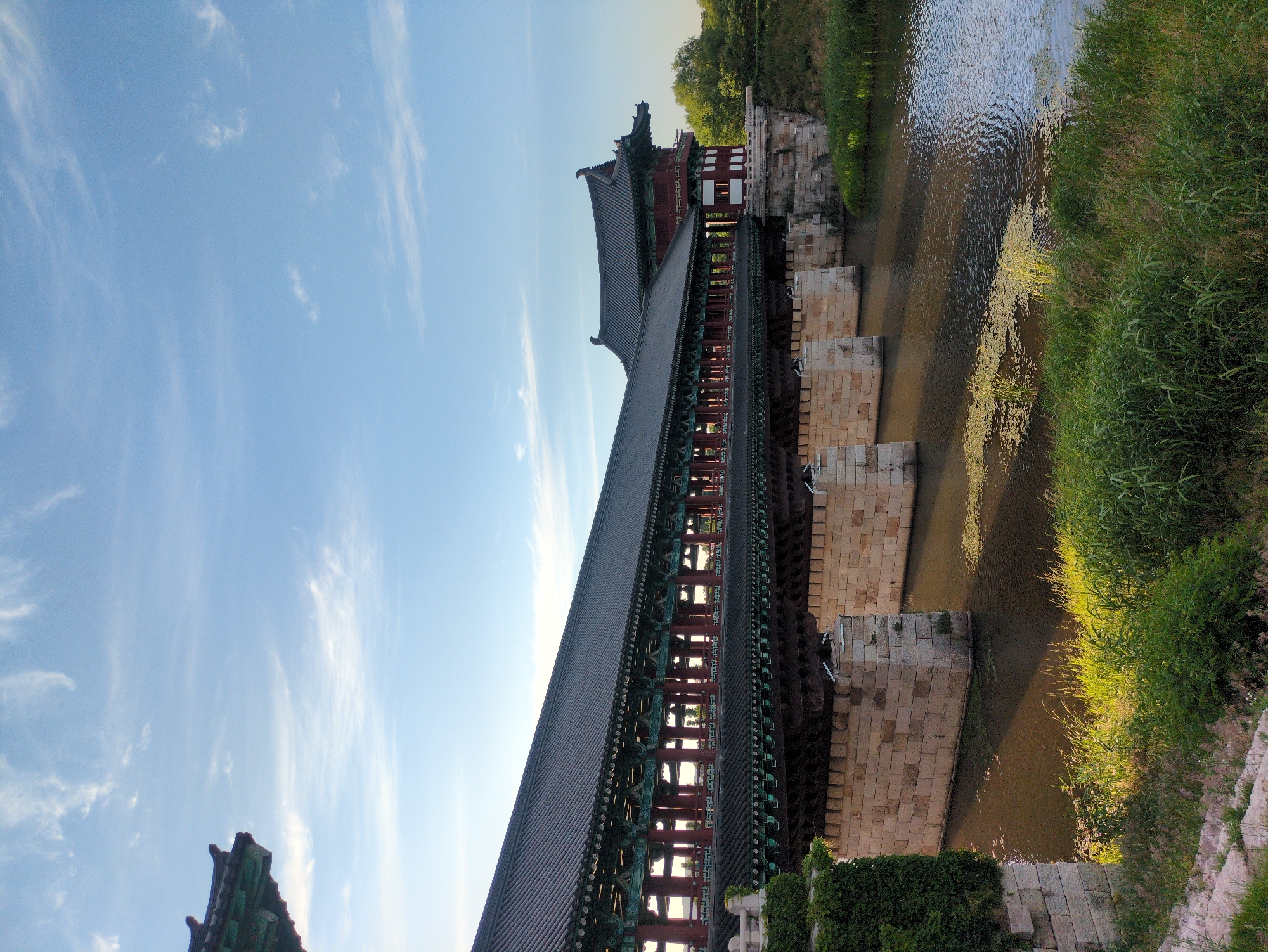 The image depicts a long, covered bridge, known as Woljeonggyo Bridge, spanning a narrow river in Gyeongju-si, South Korea. The bridge features traditional Korean architecture, with a dark gray tiled roof and red and green ornamental details along the eaves and support structures. The bridge rests on large, rectangular stone supports that rise from the riverbed. The river is shallow, with algae or vegetation visible in the water. Lush green vegetation and grass line the riverbanks. The sky above is light blue with wispy white clouds.