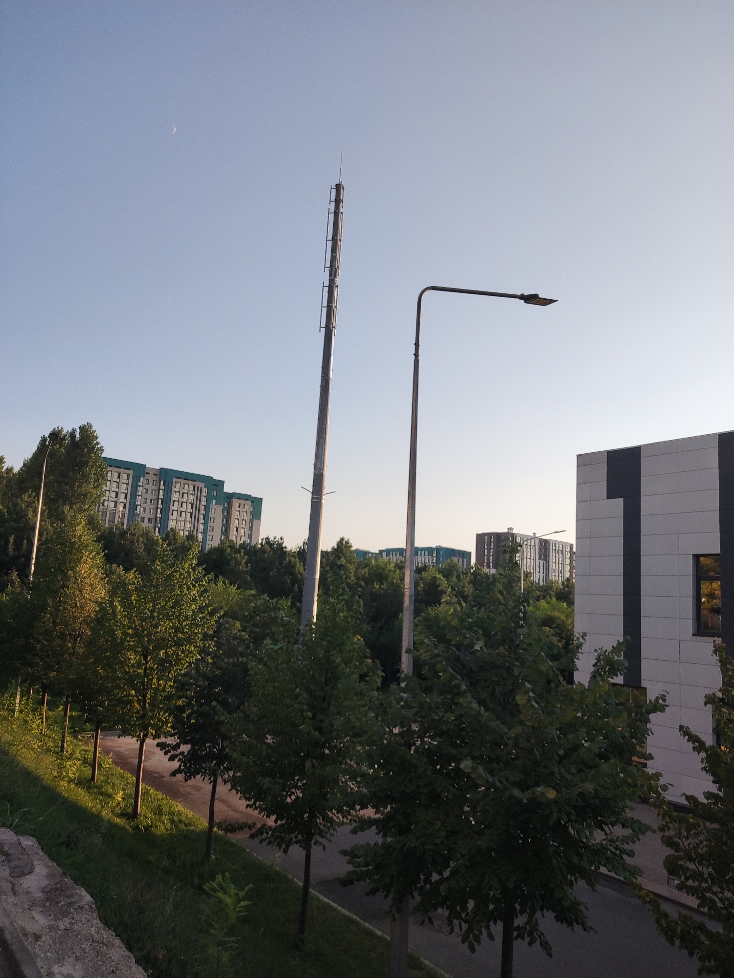 The image captures a daytime scene in the city of Shymkent, Kazakhstan. A pale crescent moon is visible in the clear blue sky. In the foreground, a row of young trees lines a road, casting long shadows due to the low angle of the sun. A concrete barrier is on the left. Behind the trees, a modern building with a light-colored facade and dark vertical accents stands on the right. Further in the background, several multi-story residential buildings with light blue and grey exteriors are visible amidst a dense canopy of green trees. Two tall metal poles, one appearing to be a communication tower with antenna-like structures, and another a street lamp, are prominent vertical elements in the composition. The overall impression is one of an urban environment with a blend of residential and commercial structures interspersed with greenery.