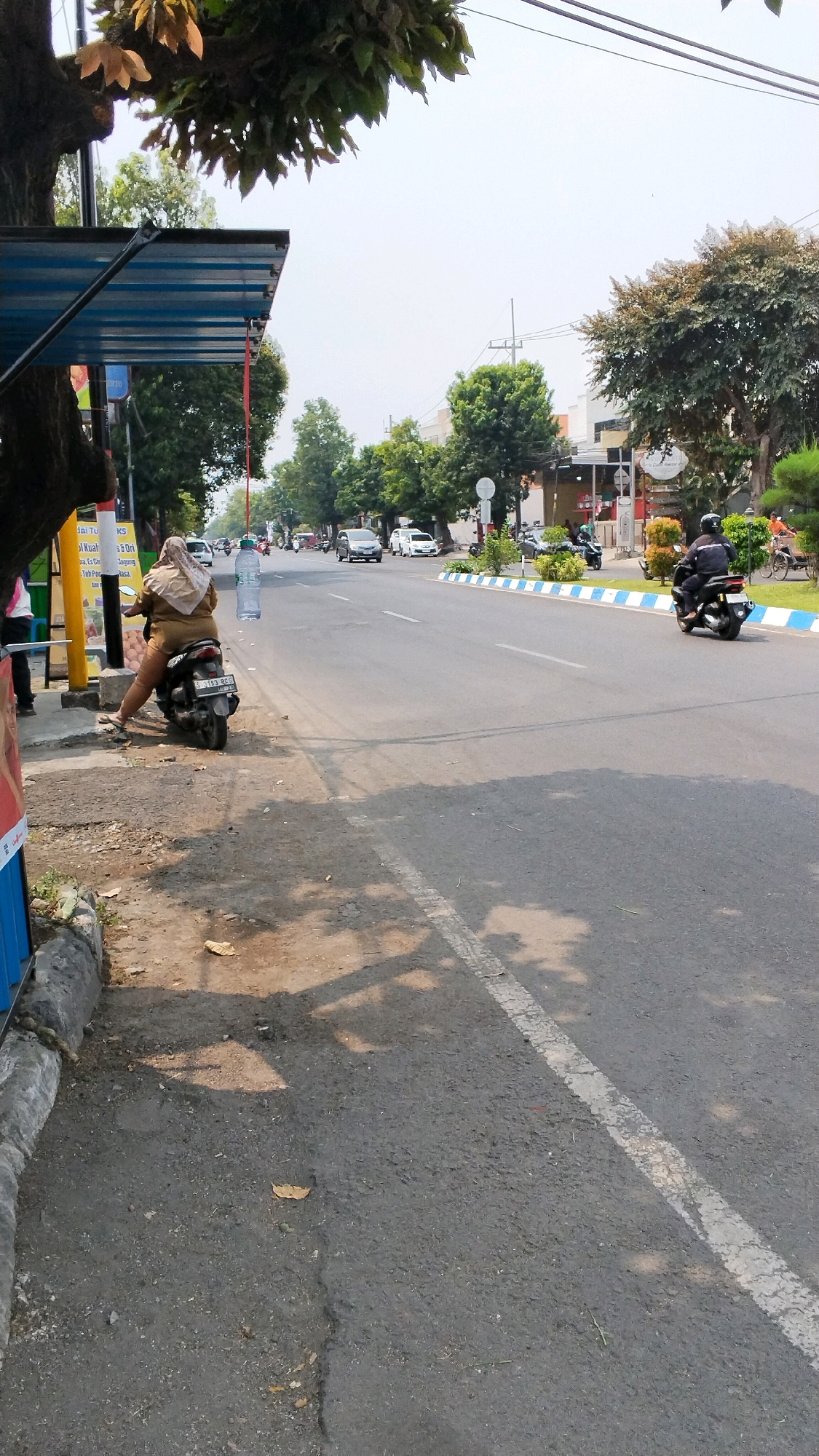 A woman in a tan hijab and matching outfit sits astride a black scooter at the edge of a paved road in Banjaragung, Indonesia. A large, clear plastic bottle of water hangs from a red string suspended in the air above and slightly in front of the scooter. The scooter's license plate reads "S 2193 CC". The woman is facing away from the camera, looking towards the road. To the left of the scooter, a blue and silver awning extends over a sidewalk area with a person in the background partially visible. Trees with green foliage line the far side of the road, with buildings visible behind them. The road is an asphalt surface with white lane markings and a blue and white striped border on the right side. Shadows from overhead trees are cast across the road and sidewalk. Several other vehicles, including cars and motorcycles, are present on the road. The sky is bright and appears to be overcast or hazy.