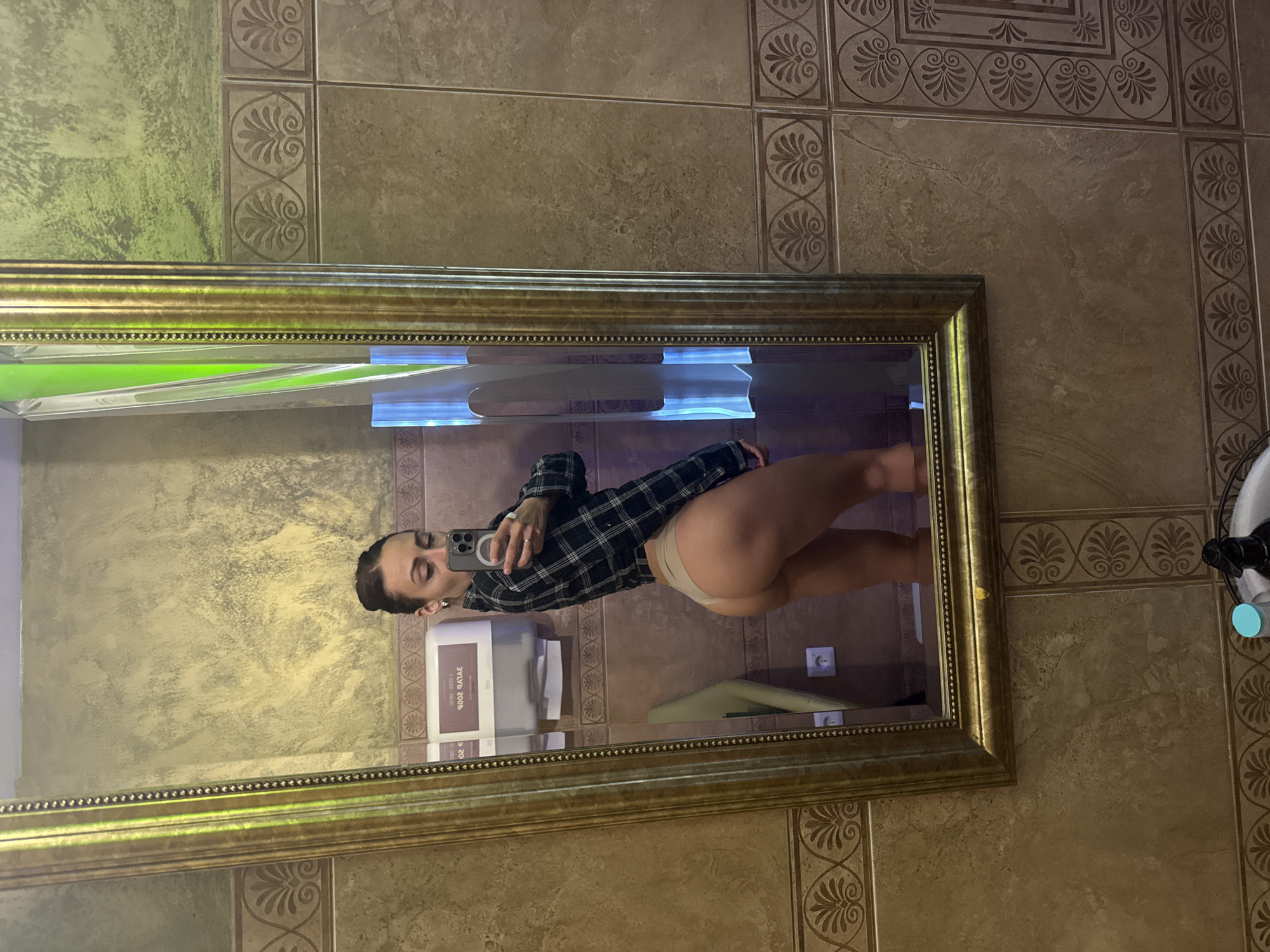 A self-portrait captured in a mirror shows a person taking a photo in what appears to be a bathroom. The image is oriented sideways, with the person standing vertically within the mirror's reflection.
The central subject is a person, likely female, reflected in a large, rectangular mirror with an ornate golden frame. She is wearing a black and white plaid long-sleeved shirt and light, possibly beige, underwear or briefs. Her dark hair is pulled back, and she is holding a dark-colored smartphone to take the picture, with her eyes squinting or closed.
The background visible in the reflection and around the mirror features tiled walls. Some tiles have a marbled, earthy brown/beige pattern, while others are highly decorative with repeating Greek key or scroll-like motifs in a slightly darker shade. Above the mirror, there's an area of wall with a textured, mottled green and gold finish.
To the left of the reflected person, a white or light gray wall-mounted dispenser is visible. Visible text on the dispenser, when read in reverse (as it's a reflection), clearly states "ЖИДКОЕ МЫЛО" (Zhidkoye Mylo), which translates to "LIQUID SOAP," and "2000ml." A brand name, possibly "TORK," is also faintly visible. Below the mirror, a light fixture emits a blue or green glow. A small, light blue object, possibly part of a faucet or plumbing, is seen on the right side.
The overall setting suggests a public restroom, potentially in a hotel, restaurant, or spa, given the decorative tiling and fixtures. The image captures a casual moment of self-documentation.