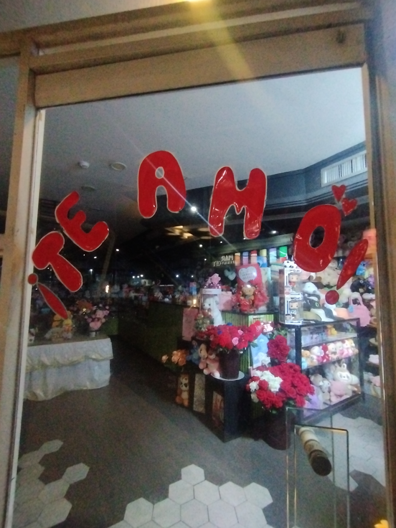 A storefront in El Valle del Espíritu Santo, Venezuela, displays a window with the words "TE AMO!" in large, red, glossy lettering. The store interior is visible through the window, revealing a variety of merchandise, including stuffed animals, flowers (mostly red roses), and displays. The floor inside is tiled with hexagonal tiles, transitioning to a darker color further in. The interior lighting is mixed, with some areas appearing brighter than others. A wooden frame surrounds the window.