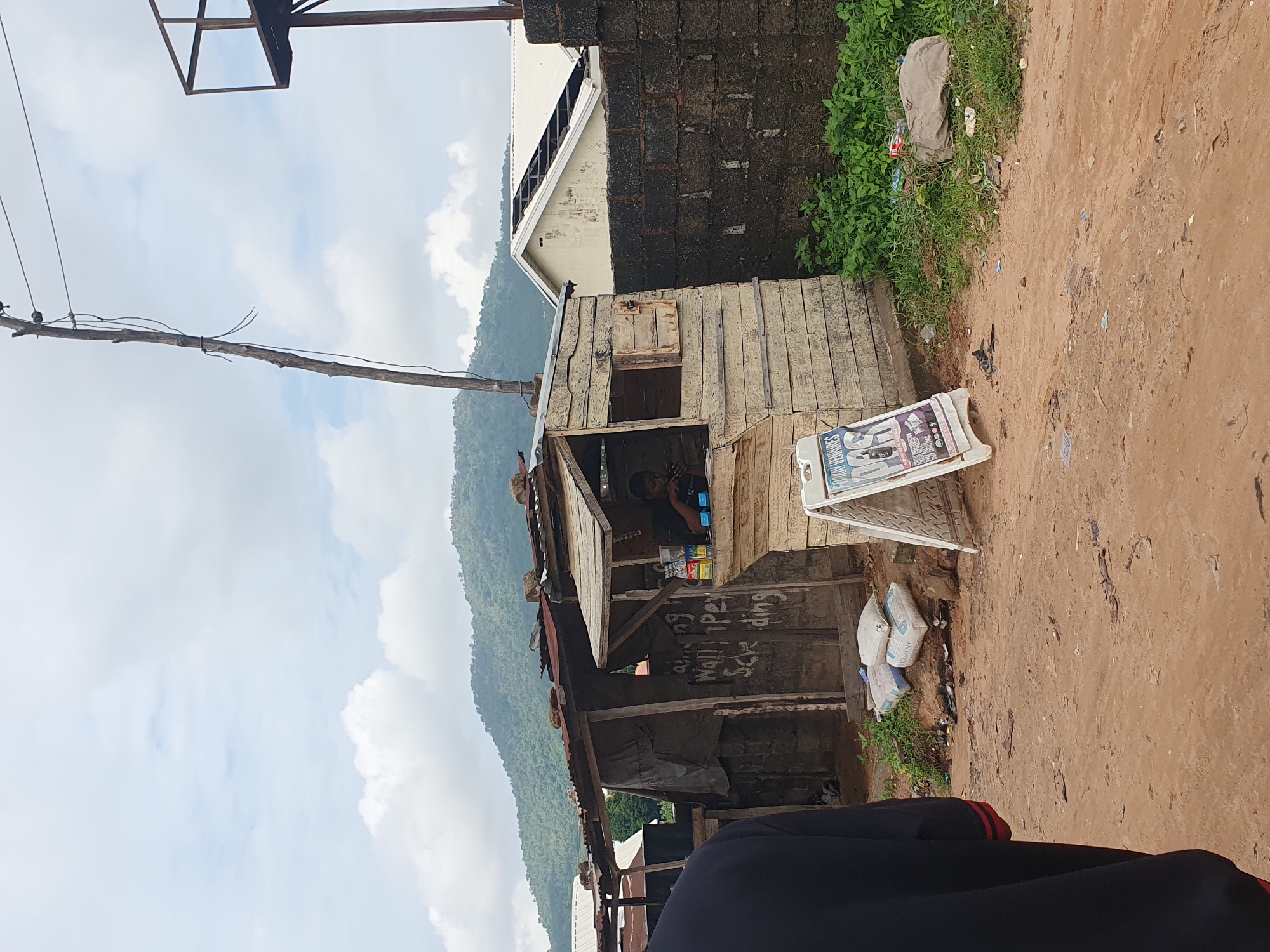 The image depicts a street scene in Lokoja, Nigeria, featuring a small, makeshift wooden stall constructed from weathered planks. The stall appears to be a retail point, with various items displayed for sale, including bags of cement, soft drinks, and possibly mobile phone accessories as suggested by an A-frame sign in front of the stall. A person is seated inside the stall, seemingly attending to business. The surrounding environment is a dusty, unpaved road with scattered debris and vegetation. In the background, a hill covered in lush green foliage rises against a cloudy sky, and a partially visible building with a damaged roof stands behind a dark brick wall. Utility poles and wires are also visible, indicating the presence of infrastructure.