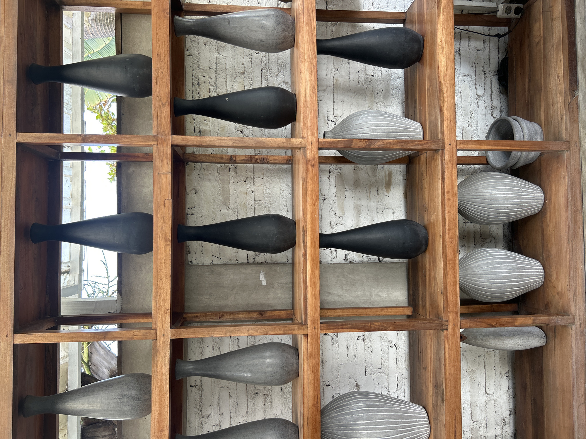 This image shows a wooden shelf unit displaying various vases and pots. The shelf unit is made of light brown wood and has multiple compartments. On the top shelf, there are two tall, slender vases, one grey and one black. The second shelf from the top also features two tall vases, one black and one grey, positioned in separate compartments. The third shelf has three items: a large ribbed grey vase on the left, a black vase in the middle, and a grey ribbed vase on the right. The bottom shelf contains a grey ribbed vase on the left, two small grey pots stacked on top of each other in the middle, and a dark grey pot on the right. The background appears to be a white brick wall. The scene is indoors, with some natural light coming from behind the shelves, suggesting an open-air or well-lit interior space. The overall aesthetic suggests a decorative display of pottery. The location is identified as Mengwi, Indonesia, likely in a shop or a home.